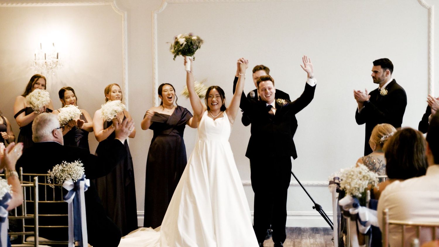 Wedding party celebrates with raised arms in a bright indoor venue with elegant decor.