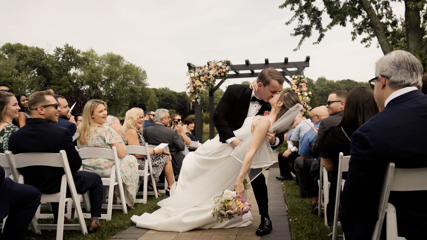 Newlyweds share a dip kiss during their outdoor ceremony as guests look on from white chairs.