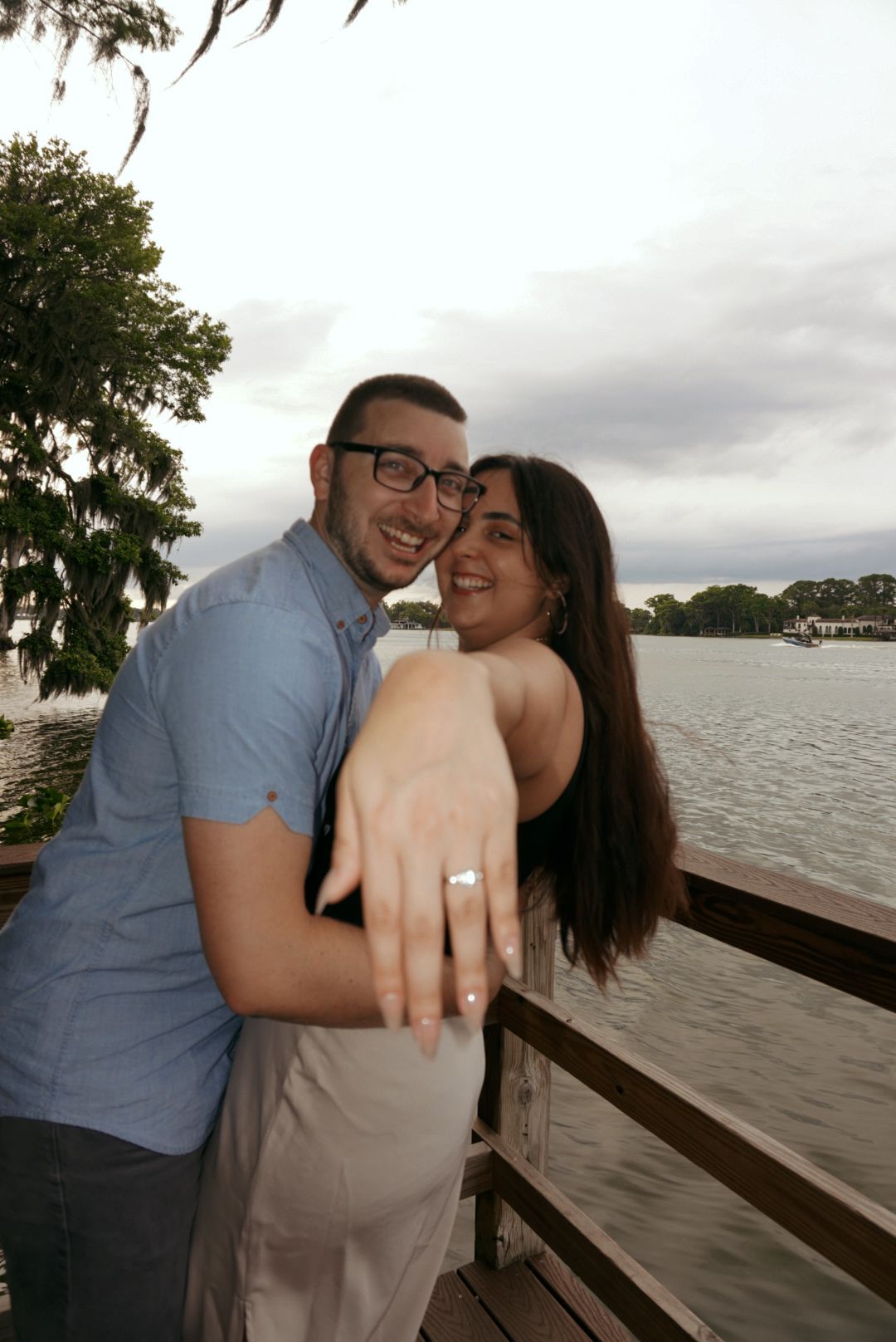 Newly engaged couple smiles and shows off diamond ring on a wooden dock by the lake.