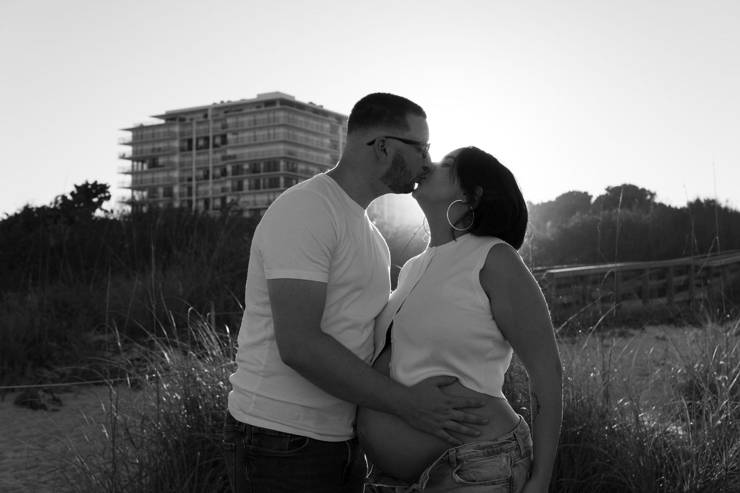 A black and white photo of a pregnant couple sharing a kiss outdoors at sunset.
