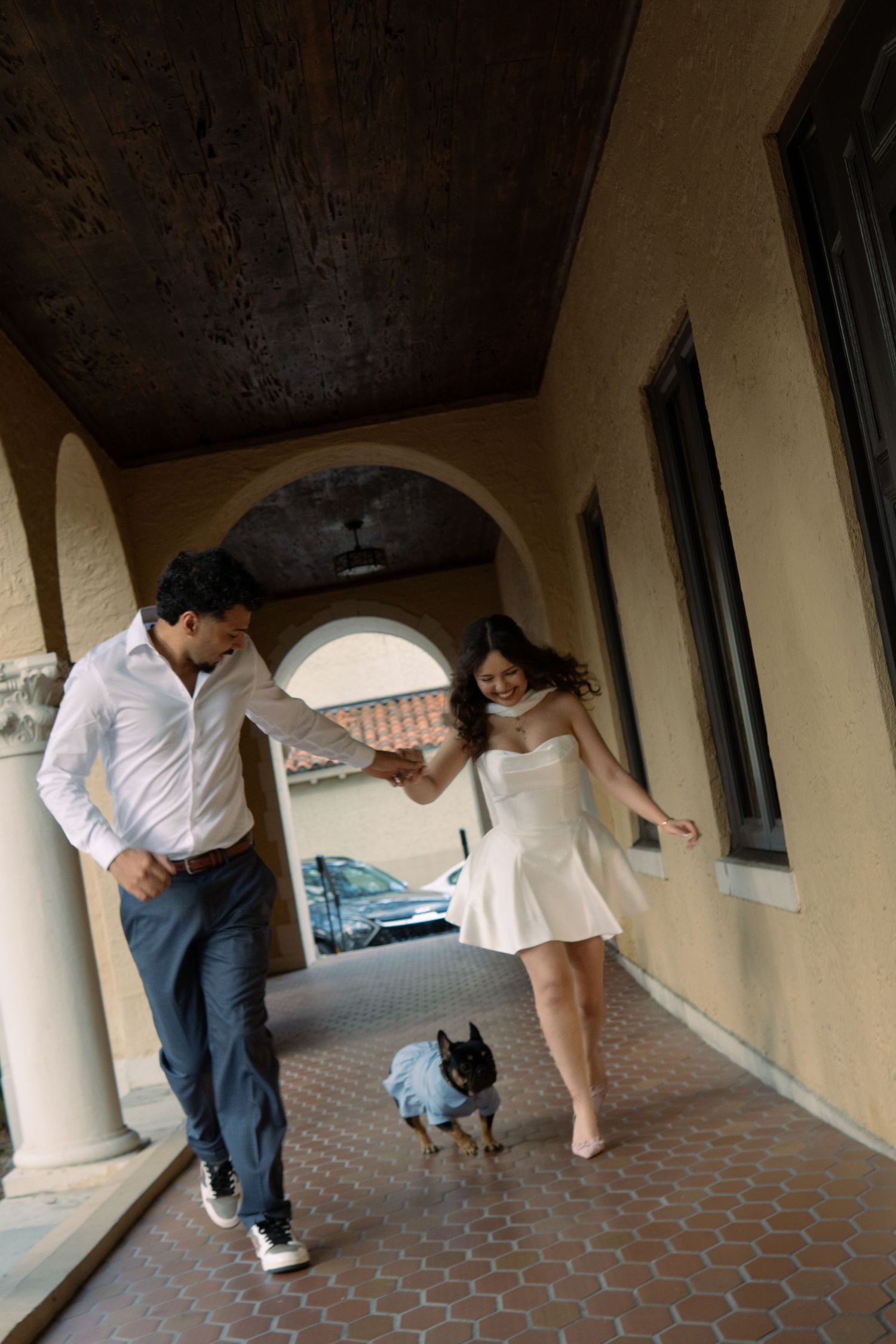 Couple walking with small dog through an arched corridor of a historic building.