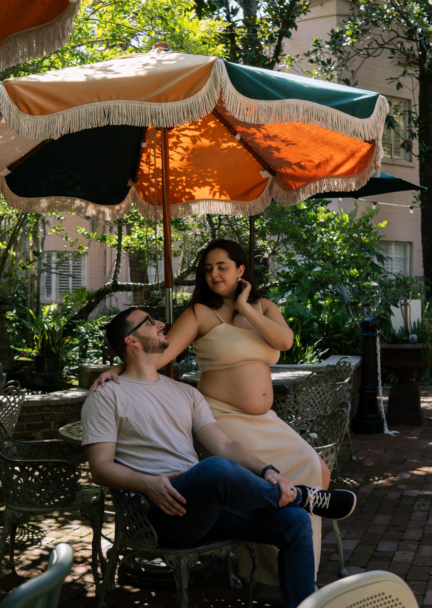 Pregnant woman in beige crop top sits with partner under colorful fringed umbrellas outdoors.