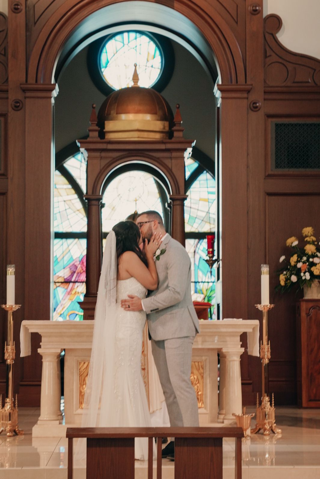 Bride and groom sharing first kiss at church altar with stained glass windows and candles in background.