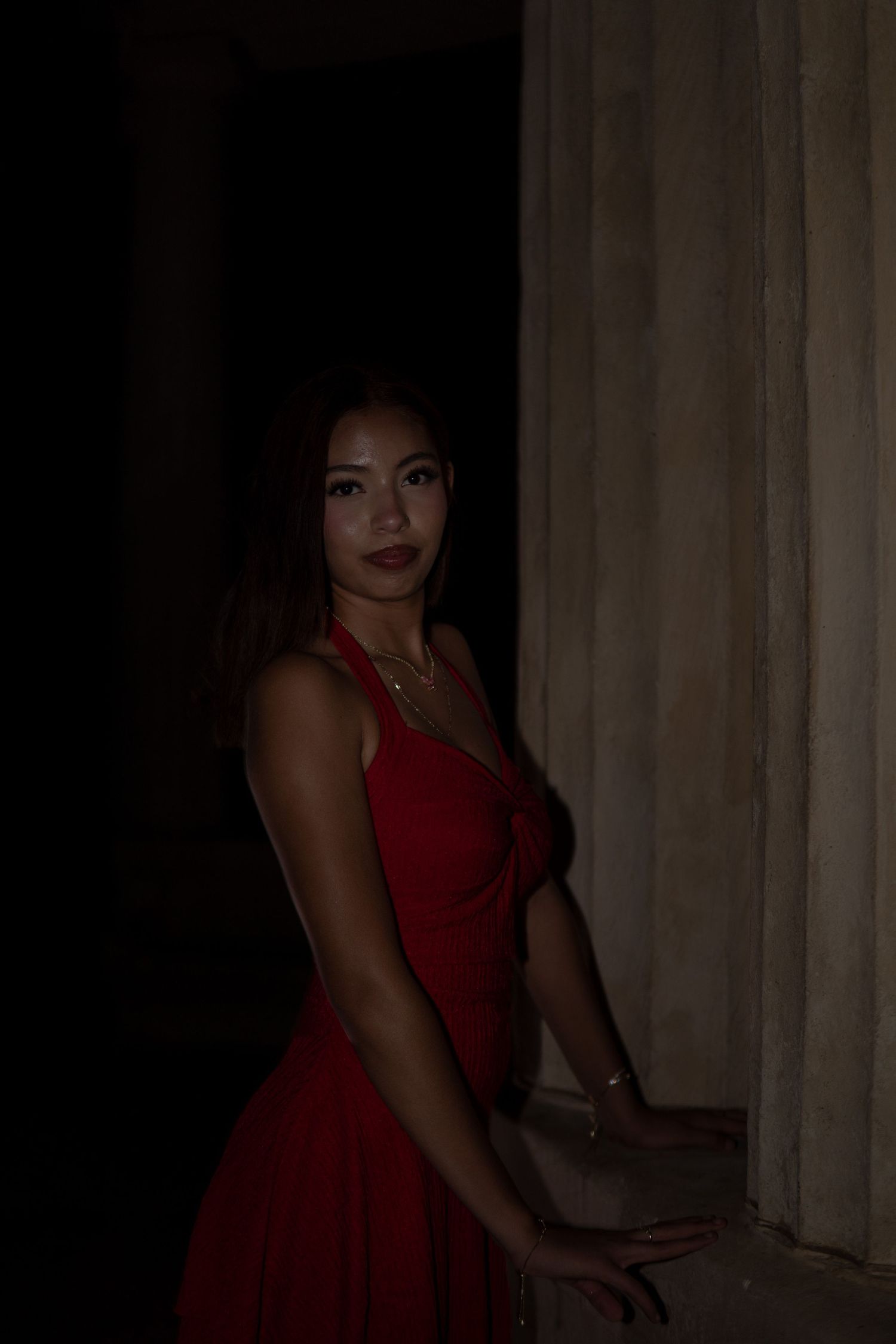 Young woman in a red halter dress posing near a stone column in dramatic low lighting.