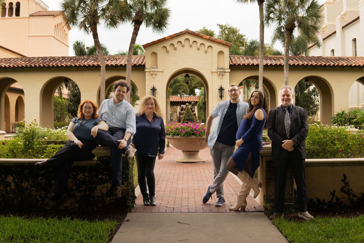 Group of seven people pose together on garden pathway in front of Mediterranean-style building.