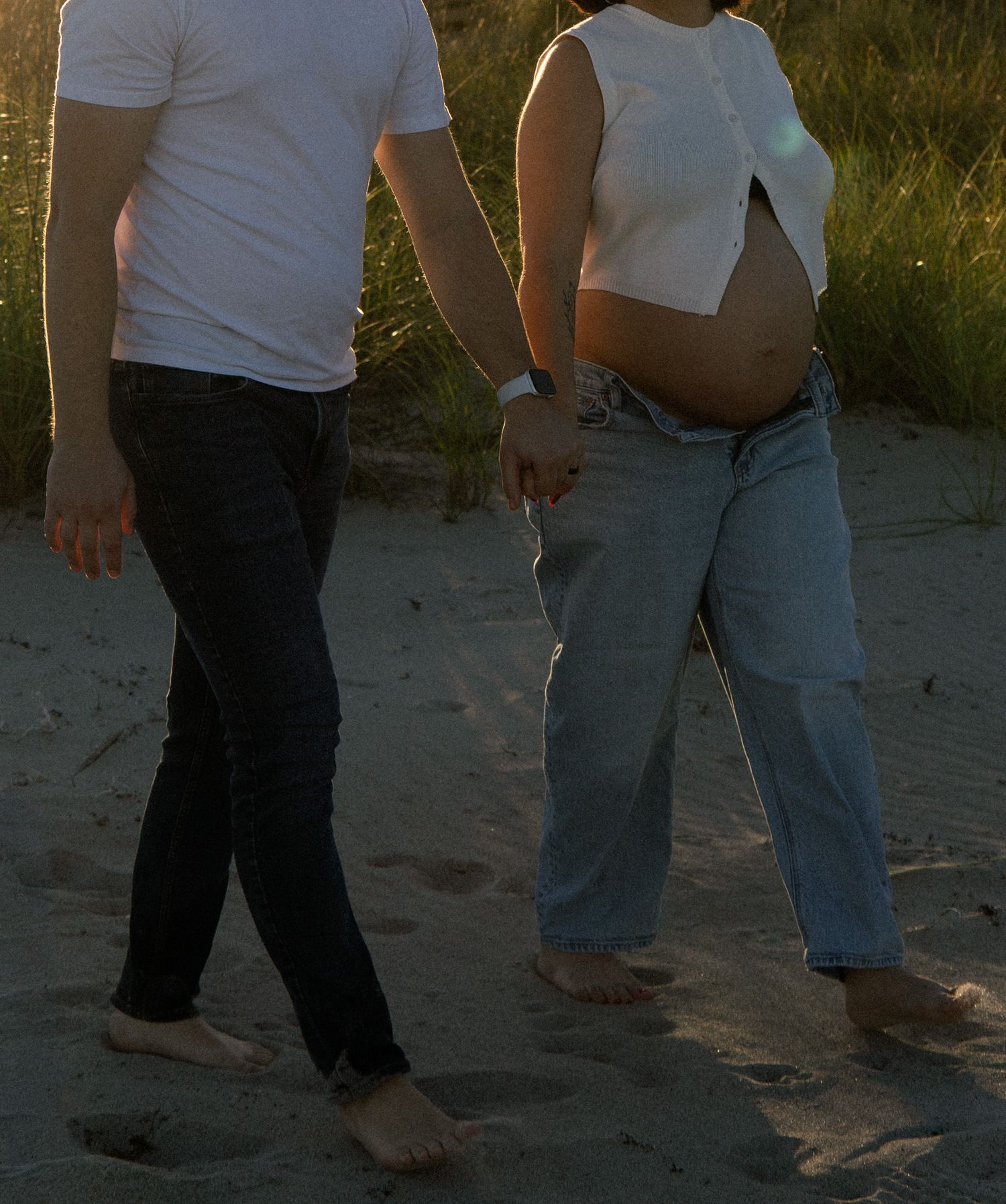 Pregnant couple walking barefoot on sandy beach, woman's baby bump visible as they hold hands at golden hour.