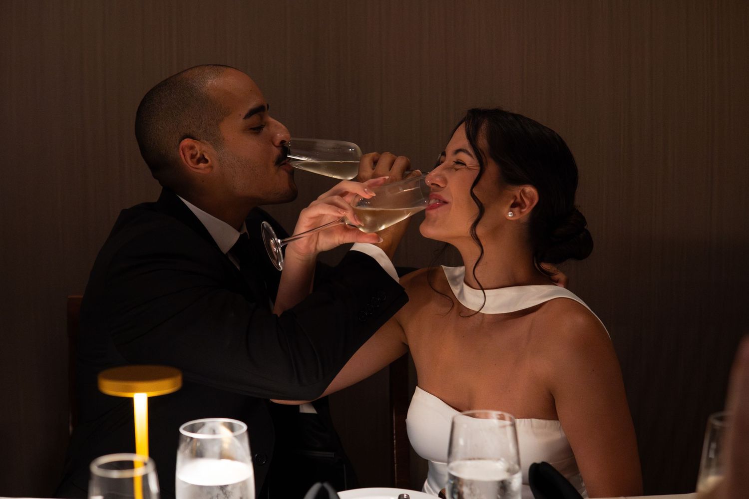 Newlywed couple sharing a champagne glass kiss at their wedding reception dinner.