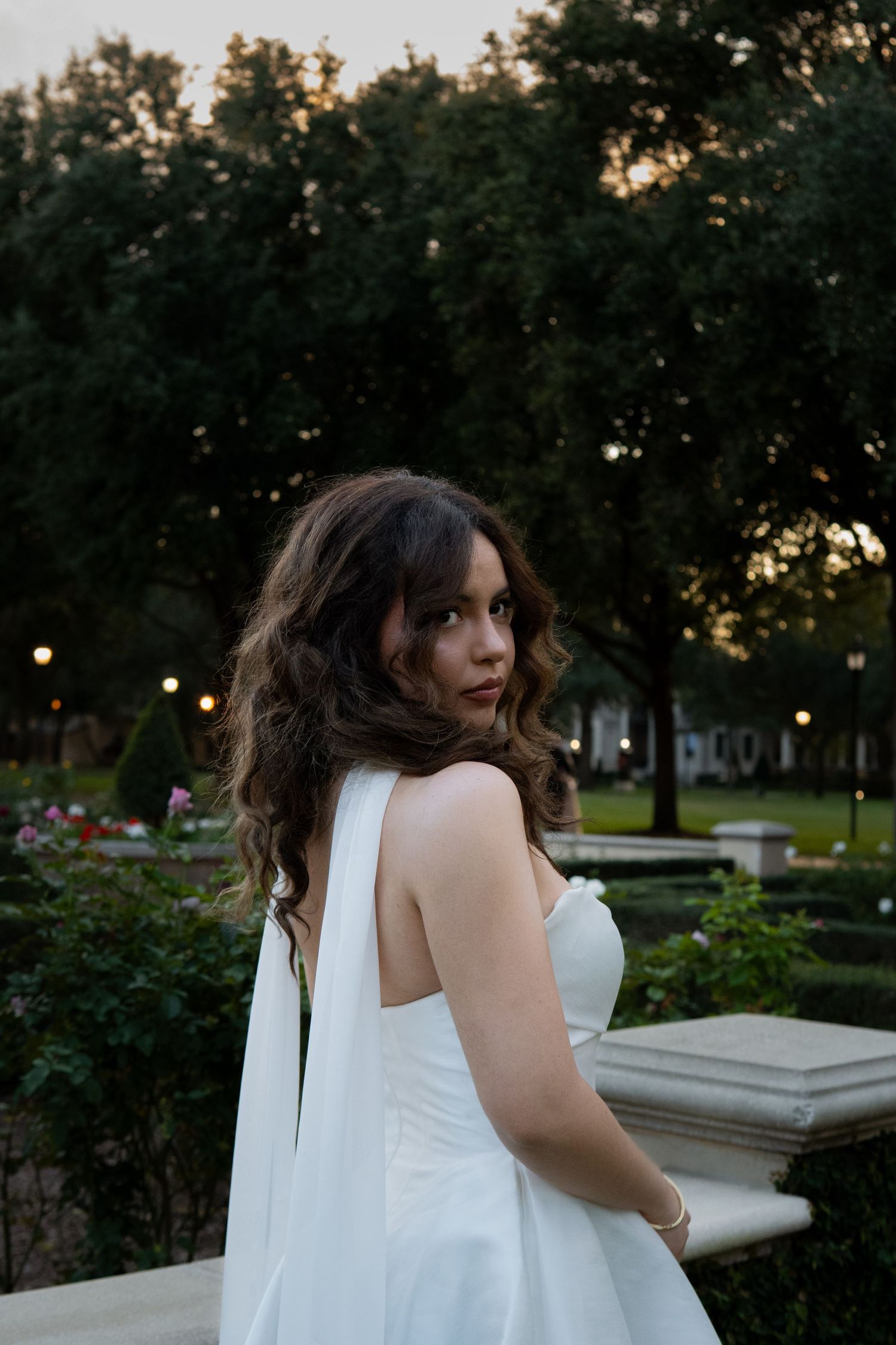 Bride in a white strapless gown with flowing cape looking back over her shoulder in an evening garden.