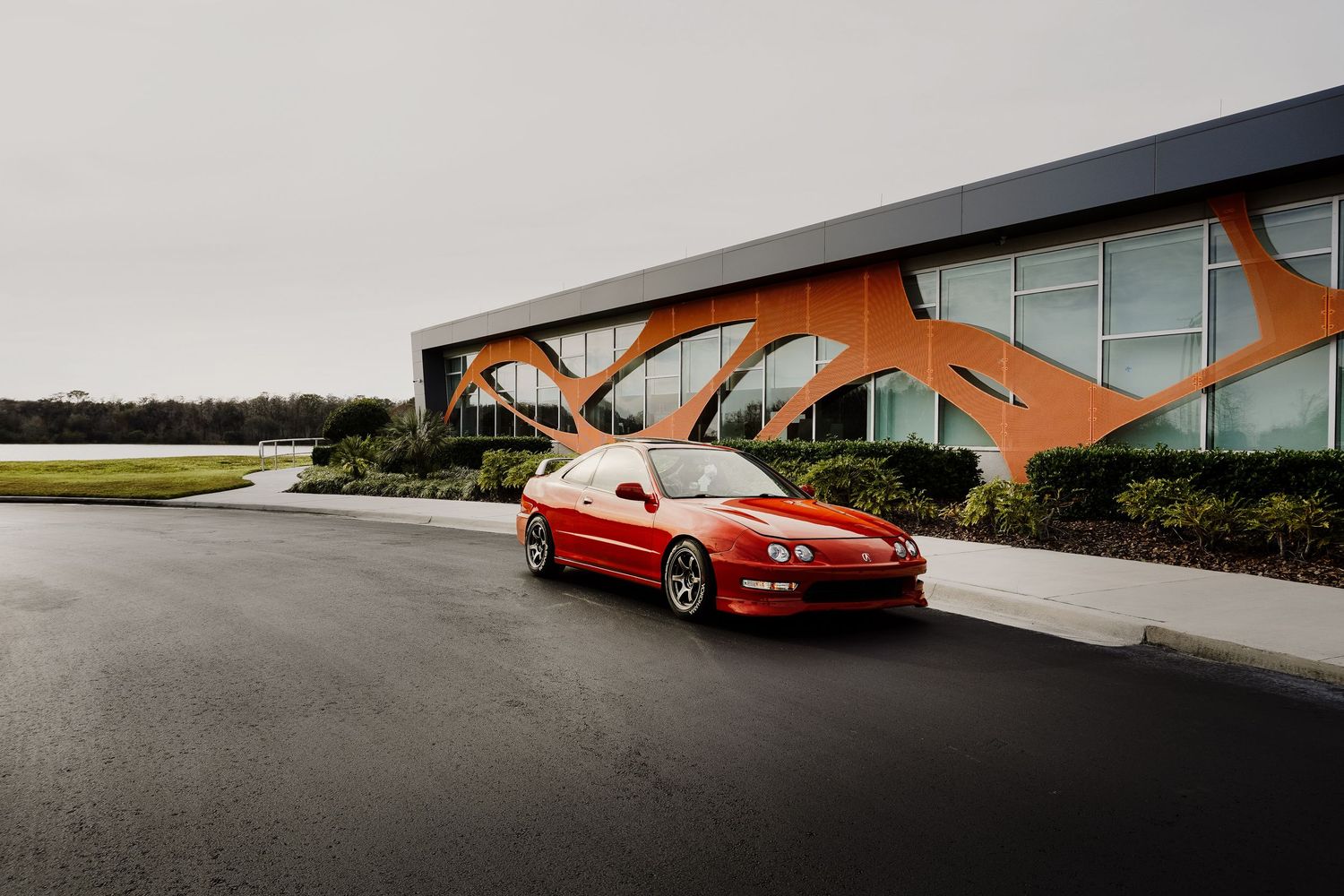 Red Acura Integra coupe parked on wet pavement in front of modern orange building.