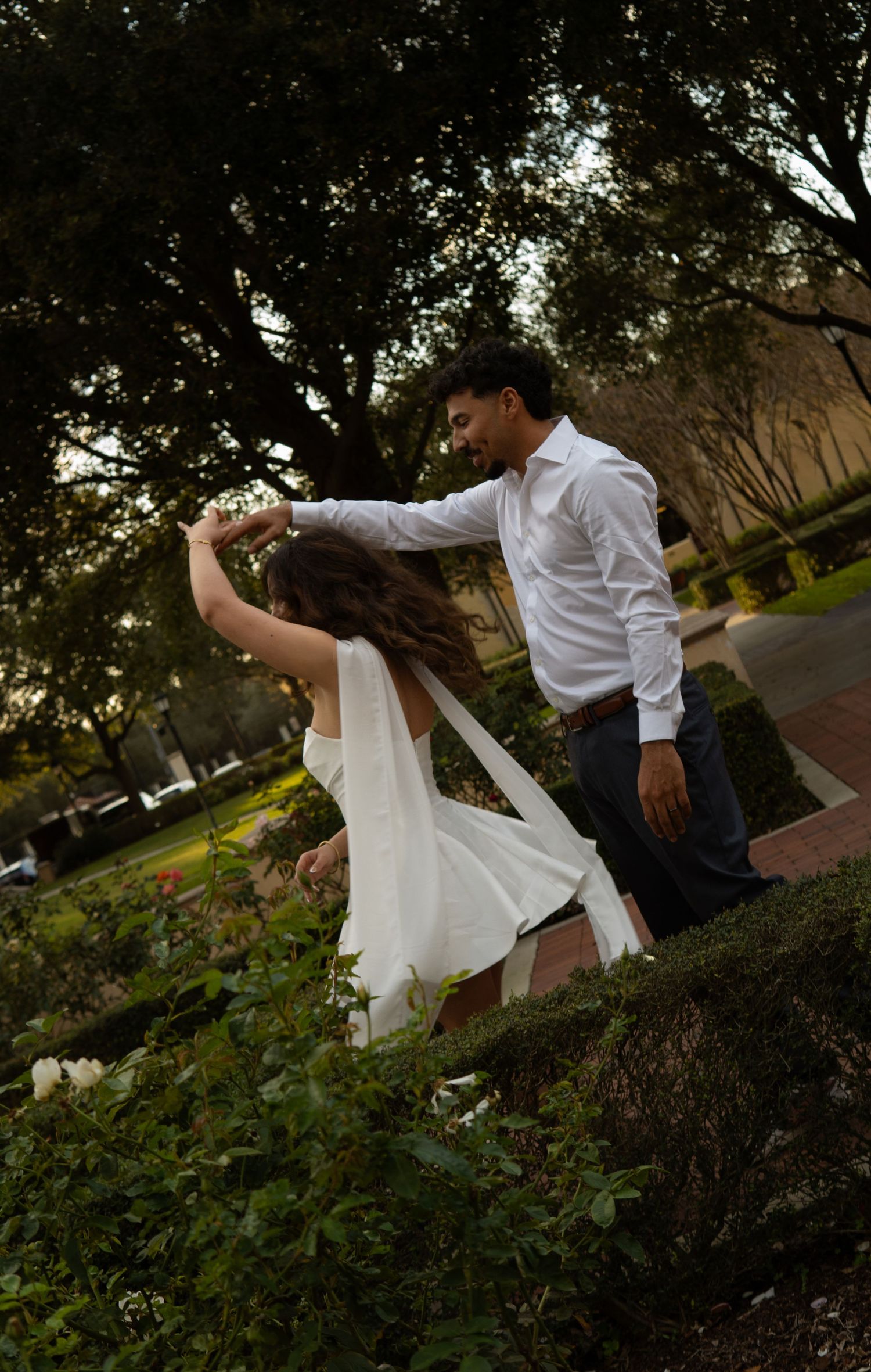 Man spinning woman in white dress outdoors near trees at golden hour.