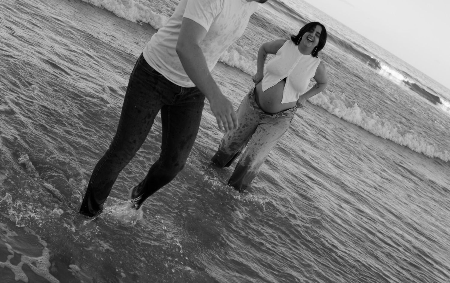 Black and white photo of pregnant couple holding hands while standing in ocean waves at the beach.