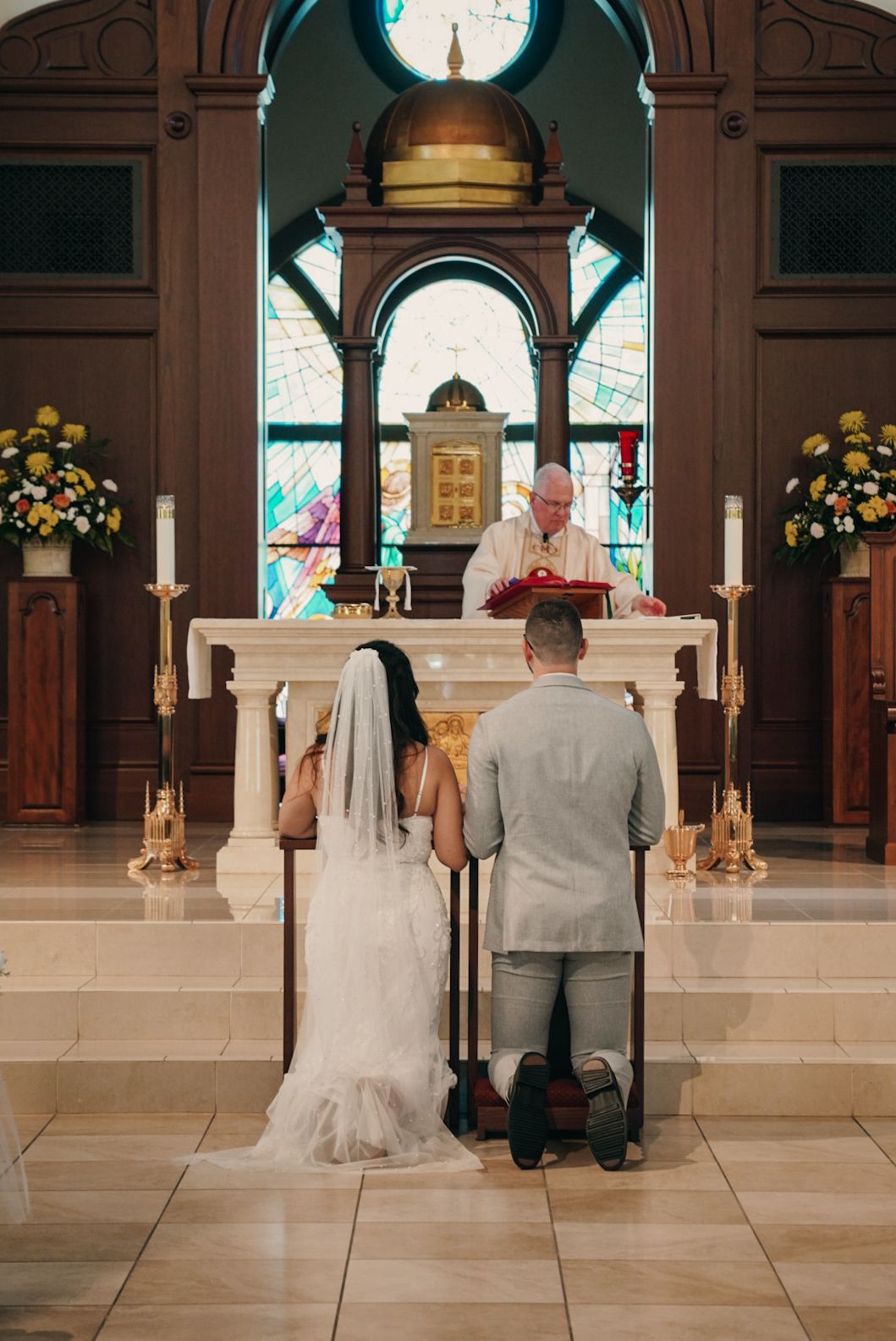 Bride and groom kneeling before priest at ornate church altar during traditional Catholic wedding ceremony.