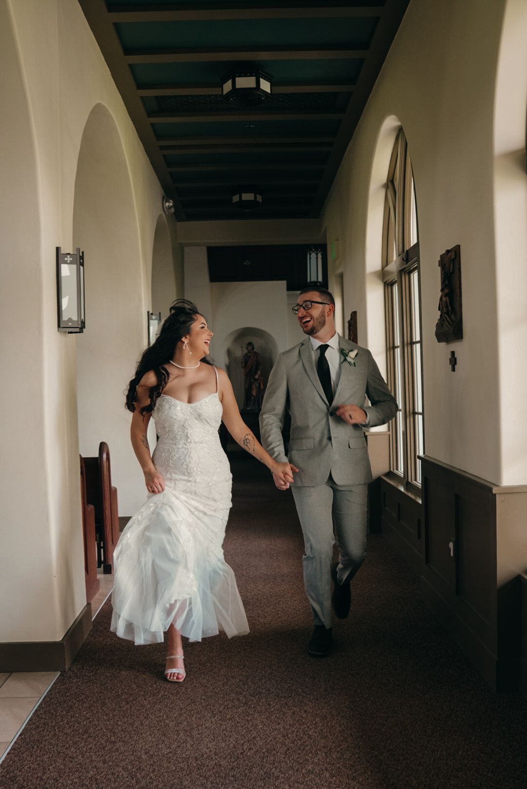 Bride in lace mermaid gown and groom in gray suit walk hand in hand through church corridor.