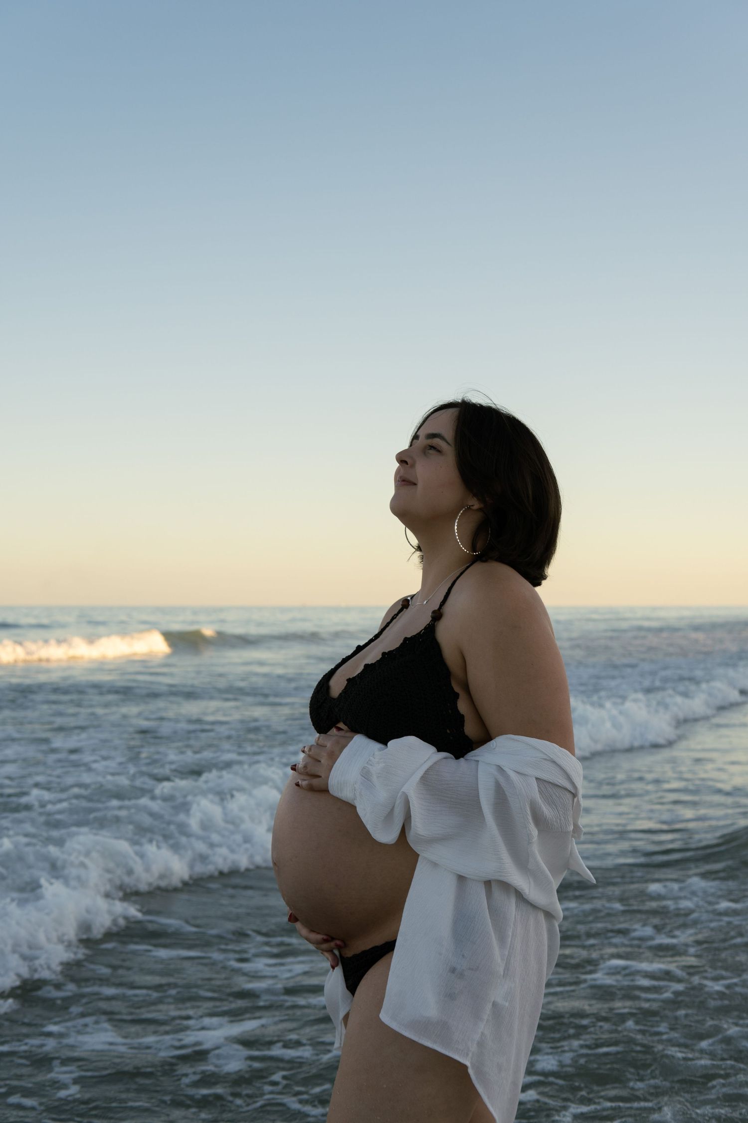 Pregnant woman in black lace bralette and open white shirt gazing upward at beach during golden hour sunset.