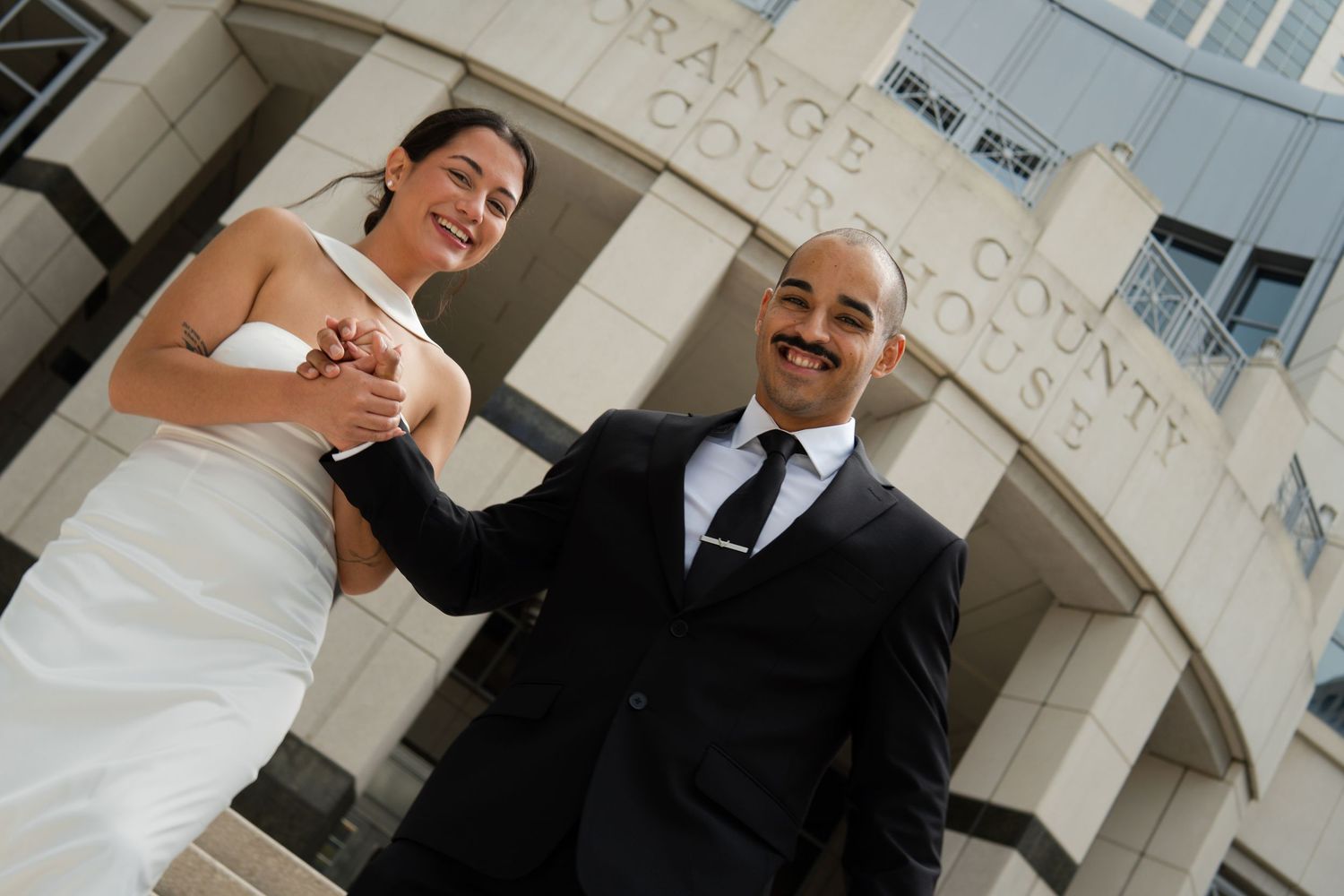 Happy newlyweds holding hands and smiling in front of Orange County Courthouse after their wedding ceremony.