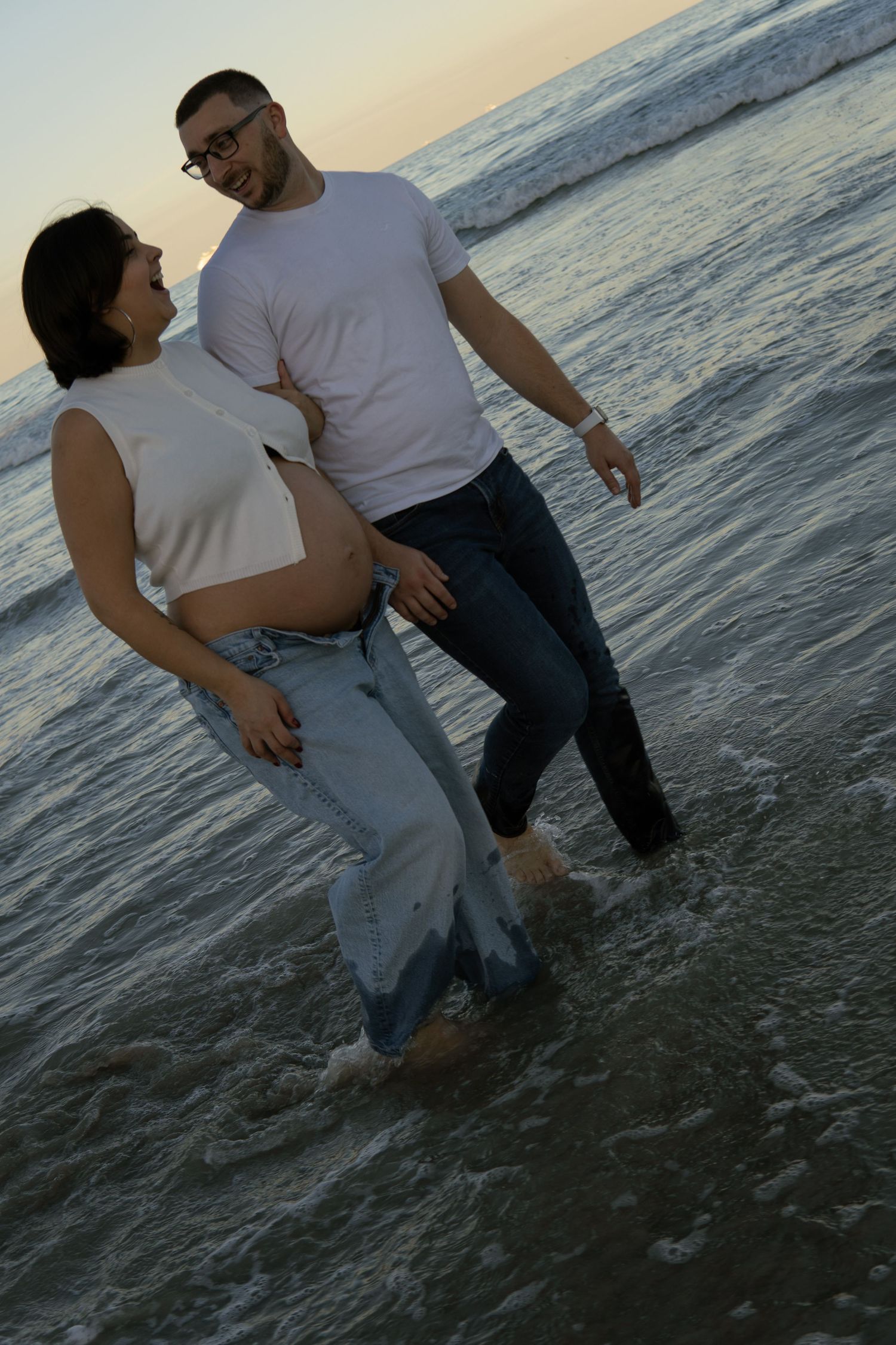 Happy pregnant couple wading in ocean waves at sunset, man smiling at partner while holding her baby bump.