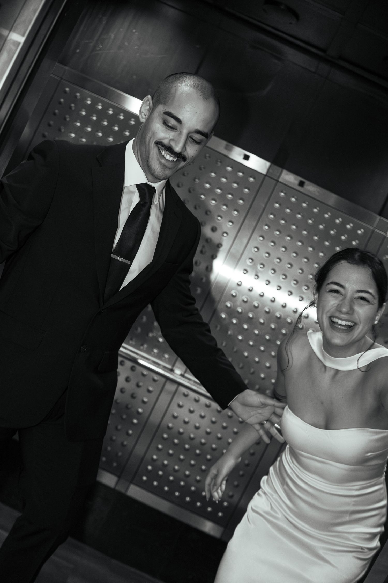 Joyful newlyweds laughing together while exiting an elevator in elegant black and white wedding attire.