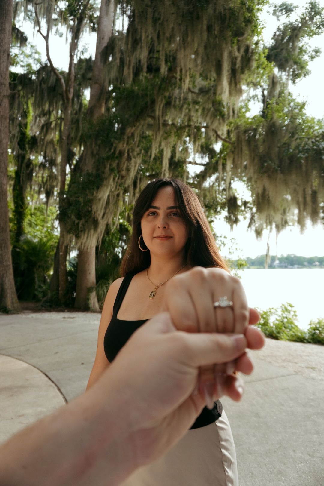 Engaged woman shows off diamond ring while holding hands near Spanish moss-draped trees by a lake.
