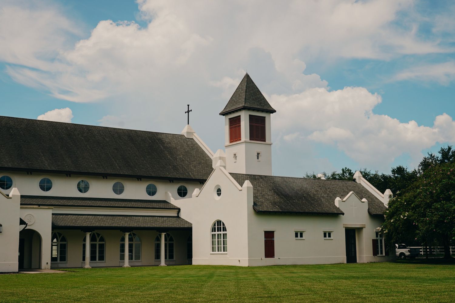 White Catholic church with bell tower, cross, and manicured lawn under partly cloudy sky.