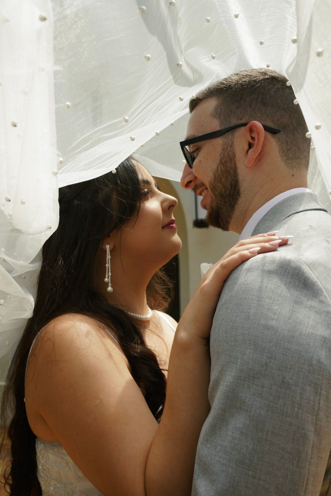 Bride and groom sharing intimate moment under white veil, groom wearing glasses.