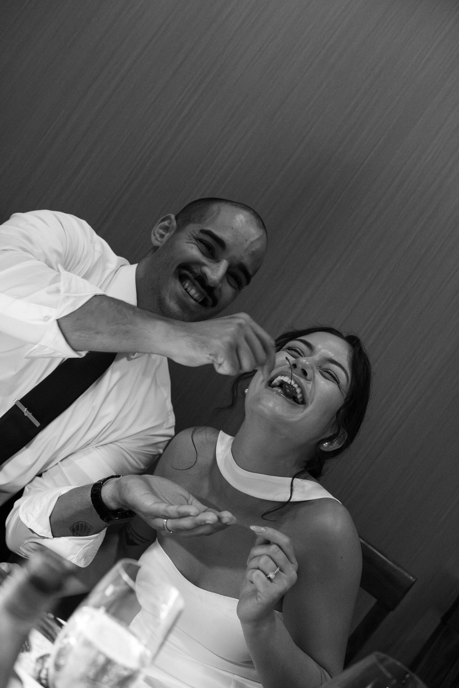 Black and white photo of laughing groom feeding cake to smiling bride at wedding reception table.