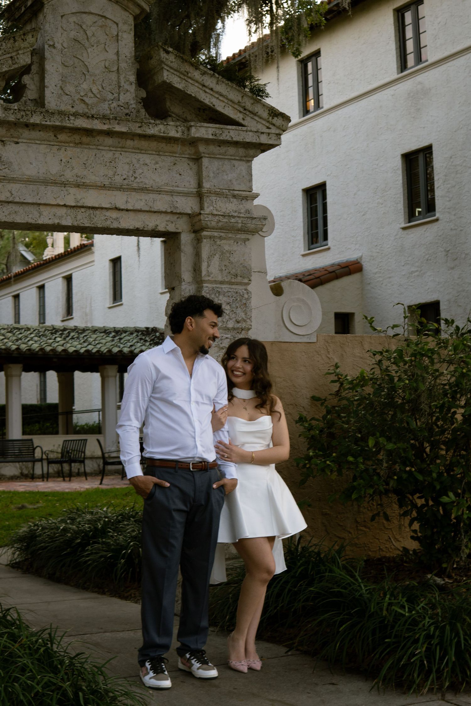 Couple in white outfits posing together outside a historic building with lush greenery.