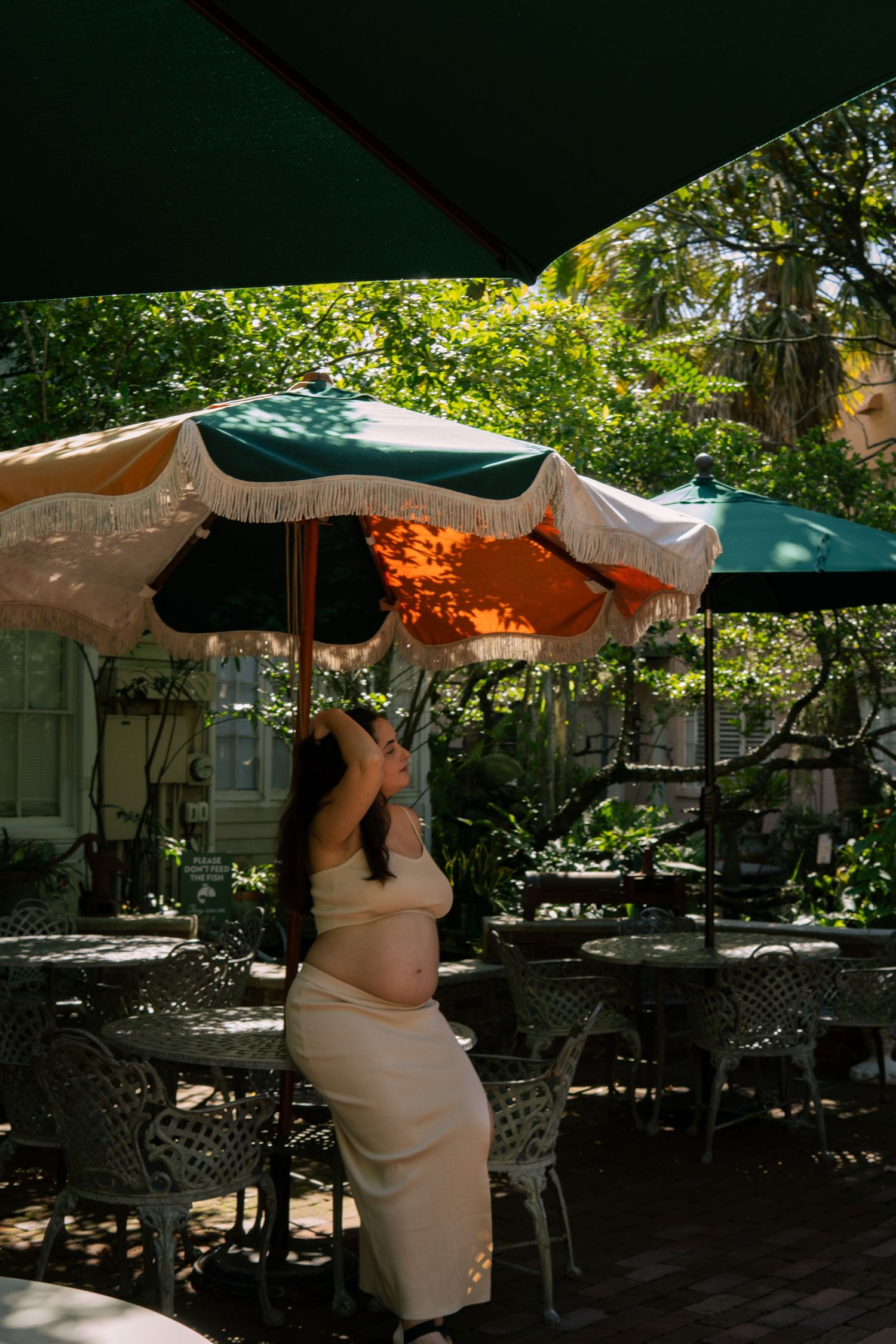 Pregnant woman in beige dress stands under colorful fringed patio umbrella in sunny garden.