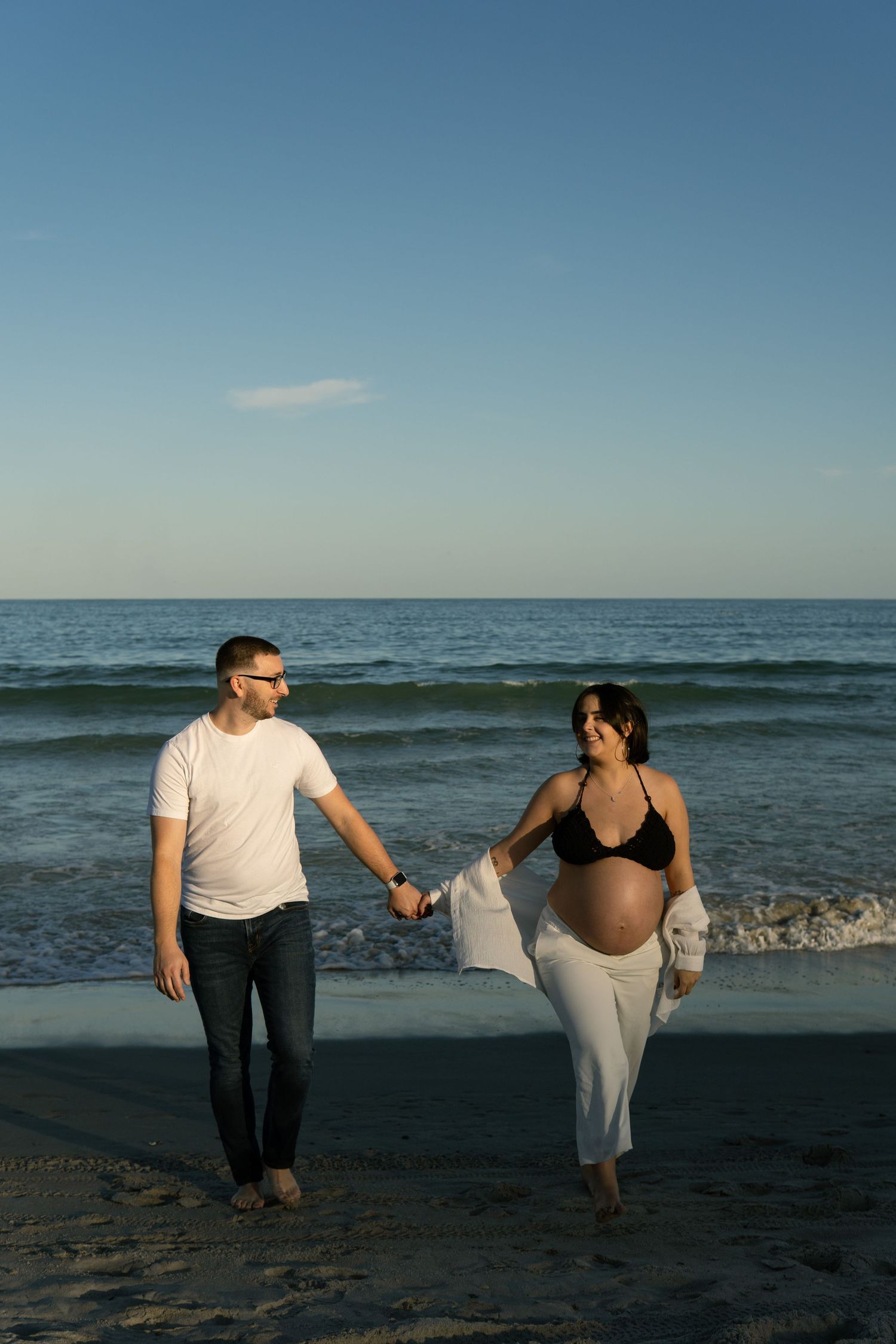 Expectant couple holds hands walking on beach at sunset, woman showing baby bump.