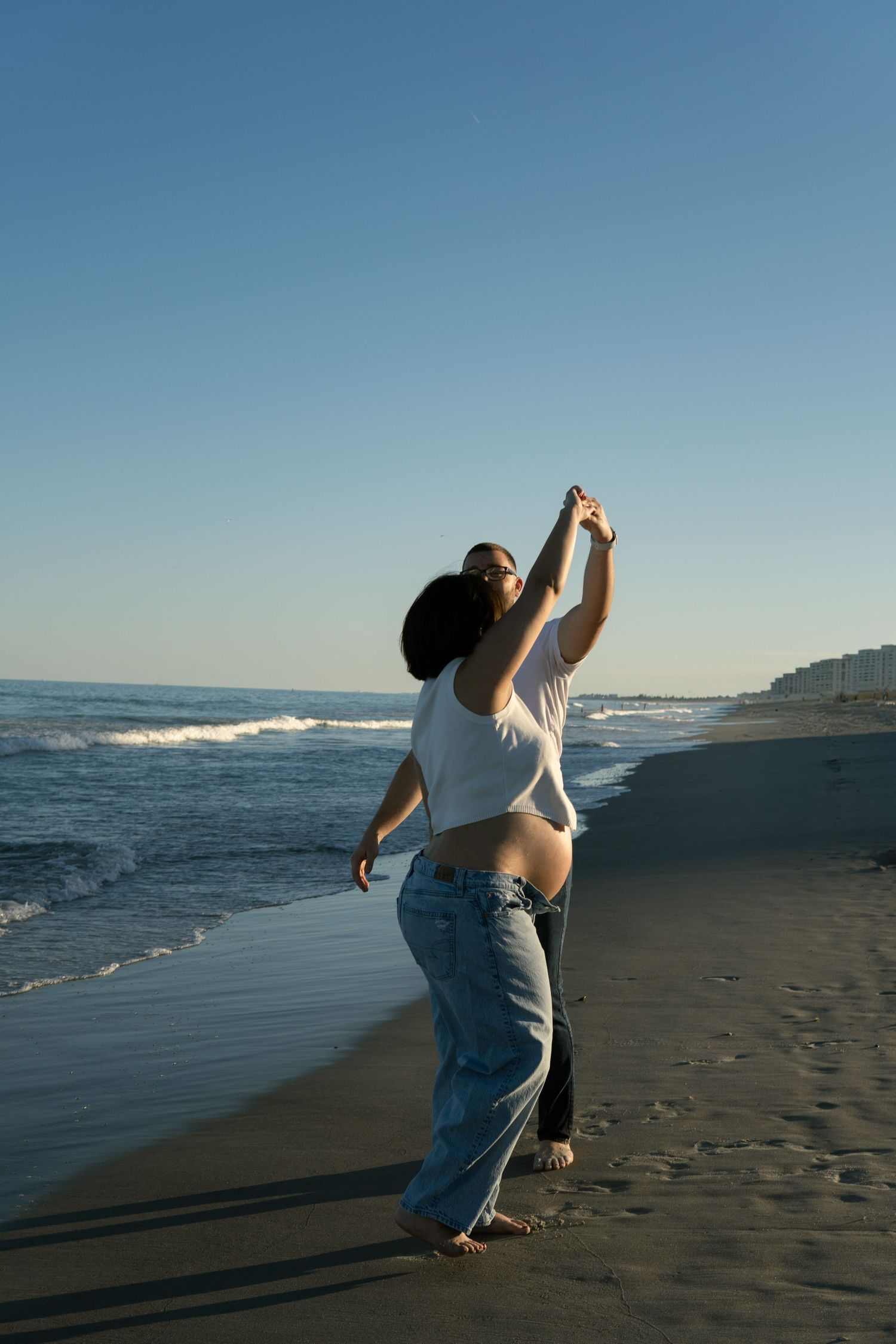 A couple dances playfully on a beach at sunset, the woman showing her pregnant belly.