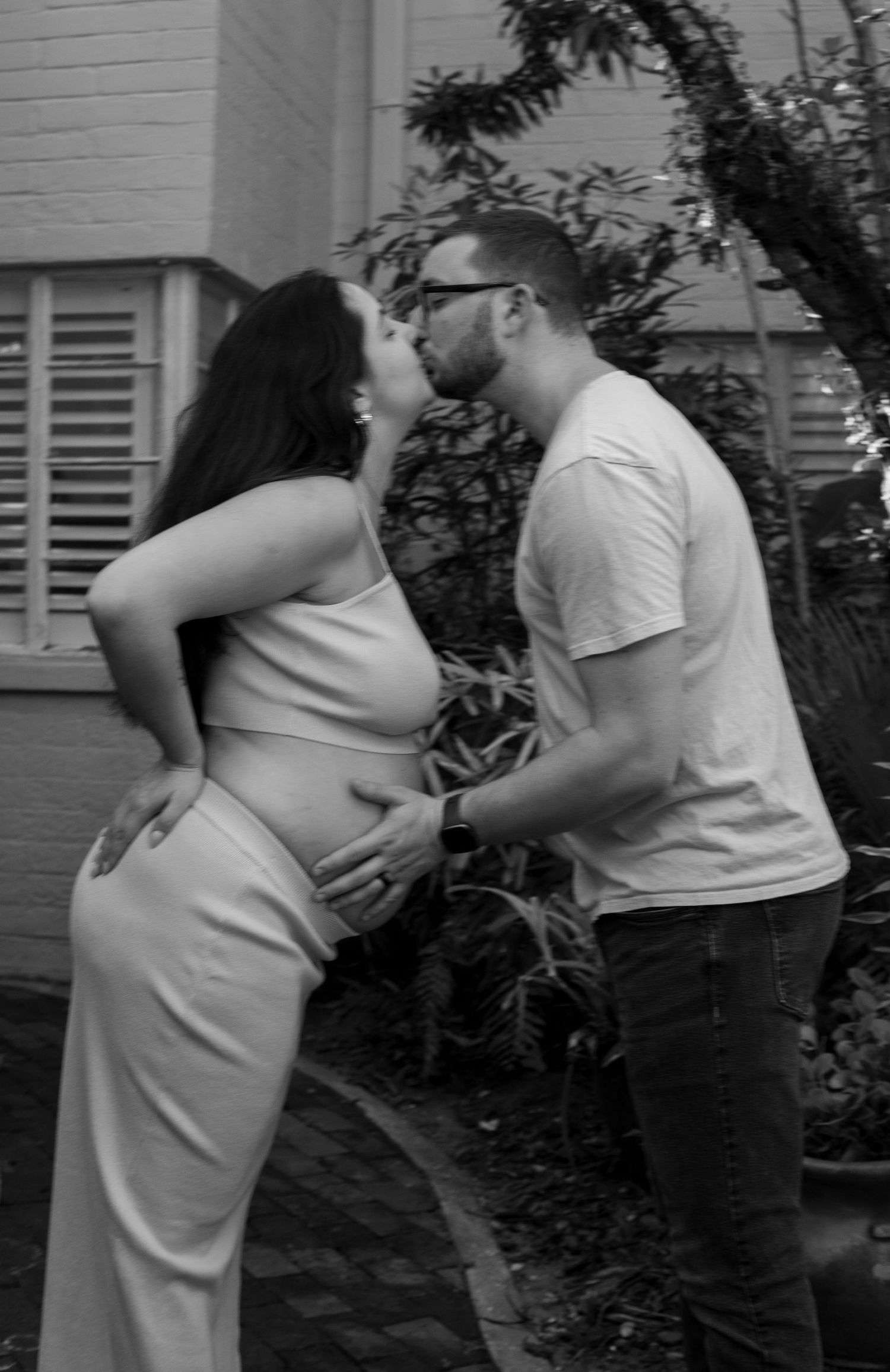 Black and white photo of pregnant couple kissing in garden, man cradling baby bump.