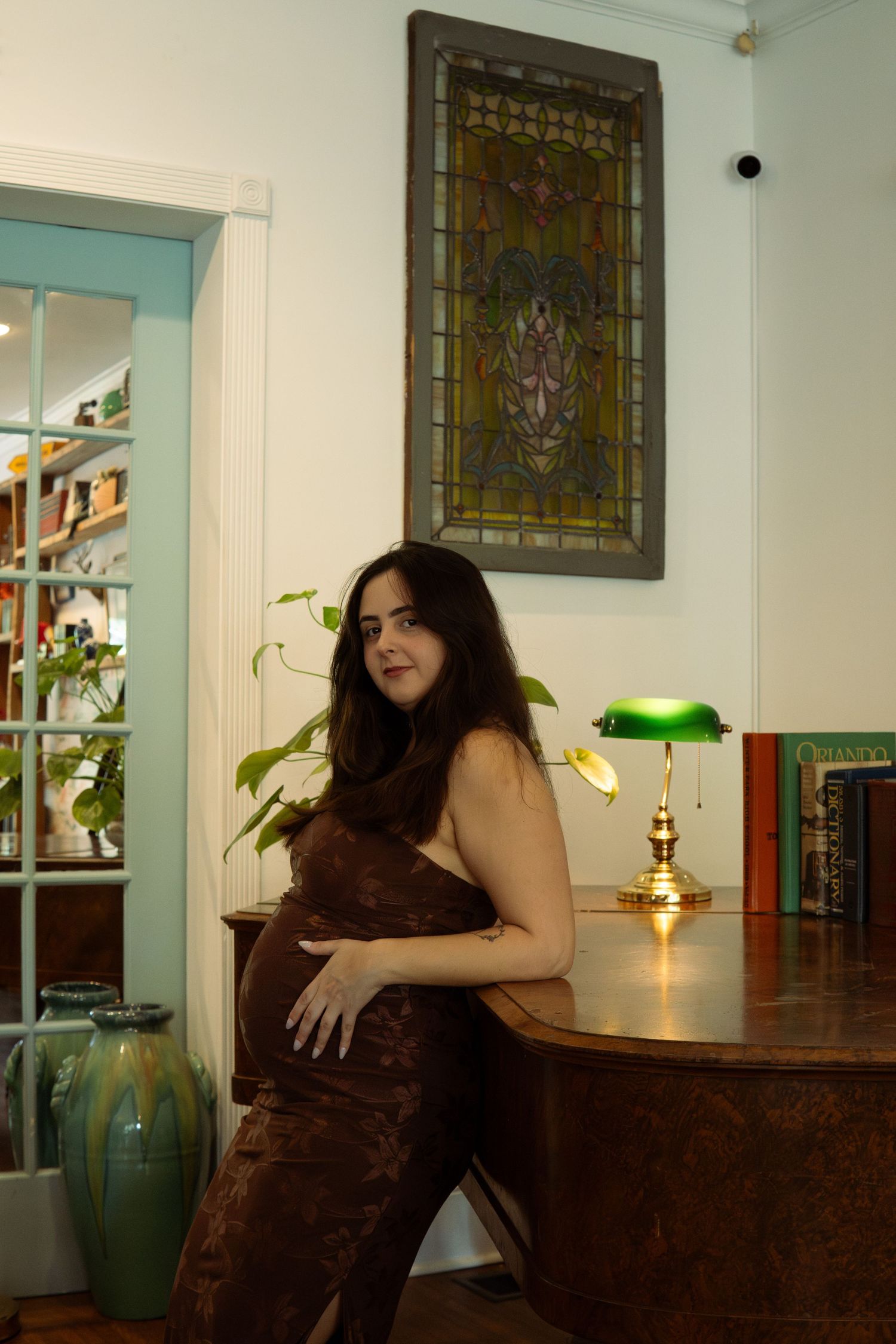 Pregnant woman in brown dress poses by antique desk with green banker lamp and stained glass window.