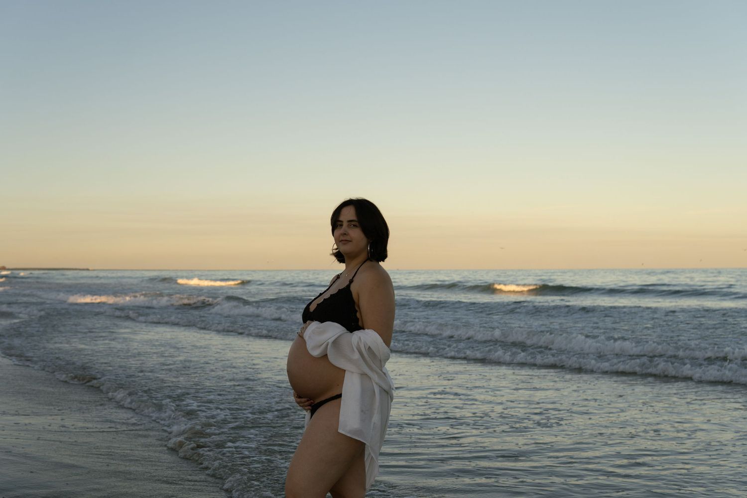 Pregnant woman in black bikini poses on beach shore during golden hour sunset.