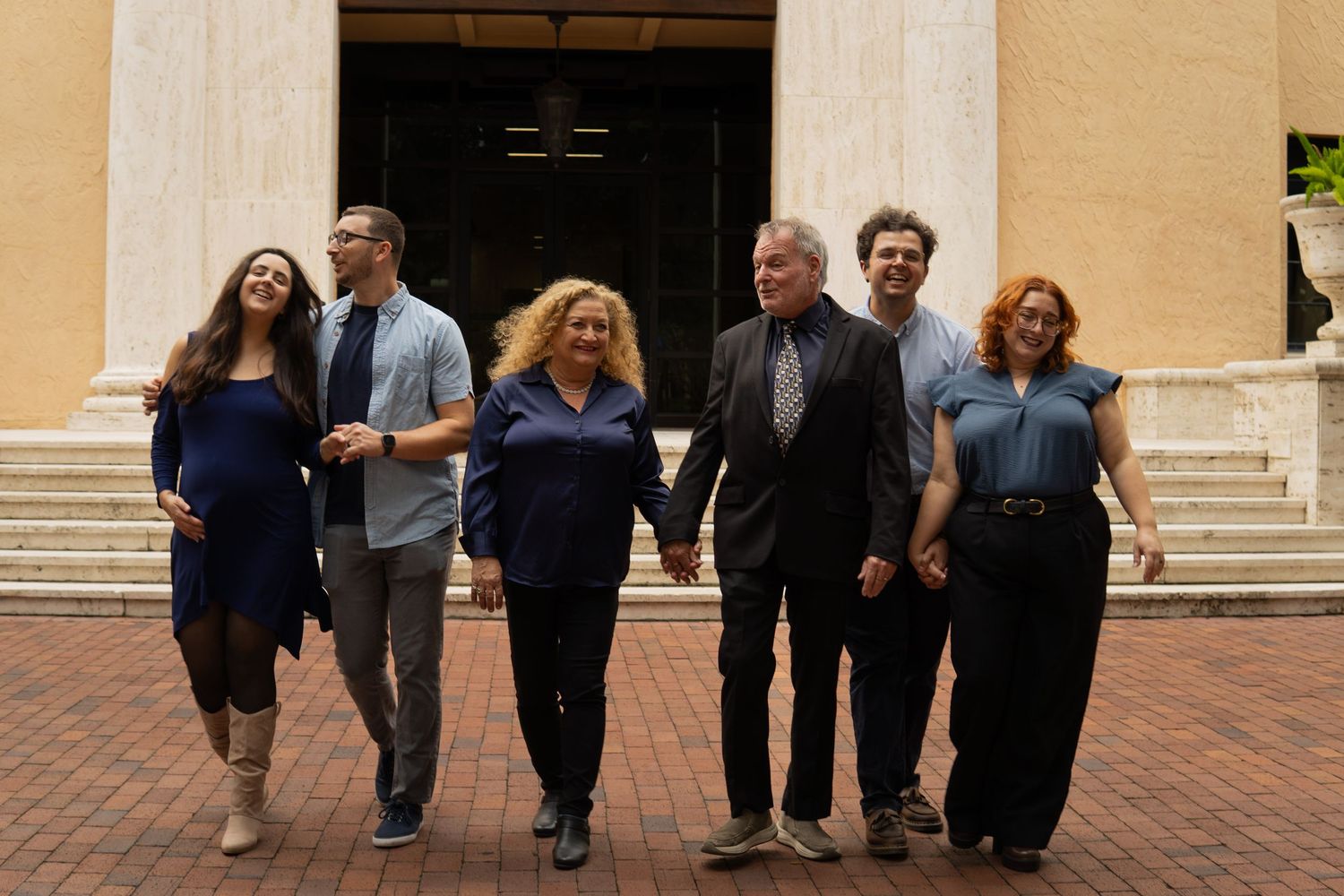 Six adults walking hand in hand along a brick path in front of a classical building.