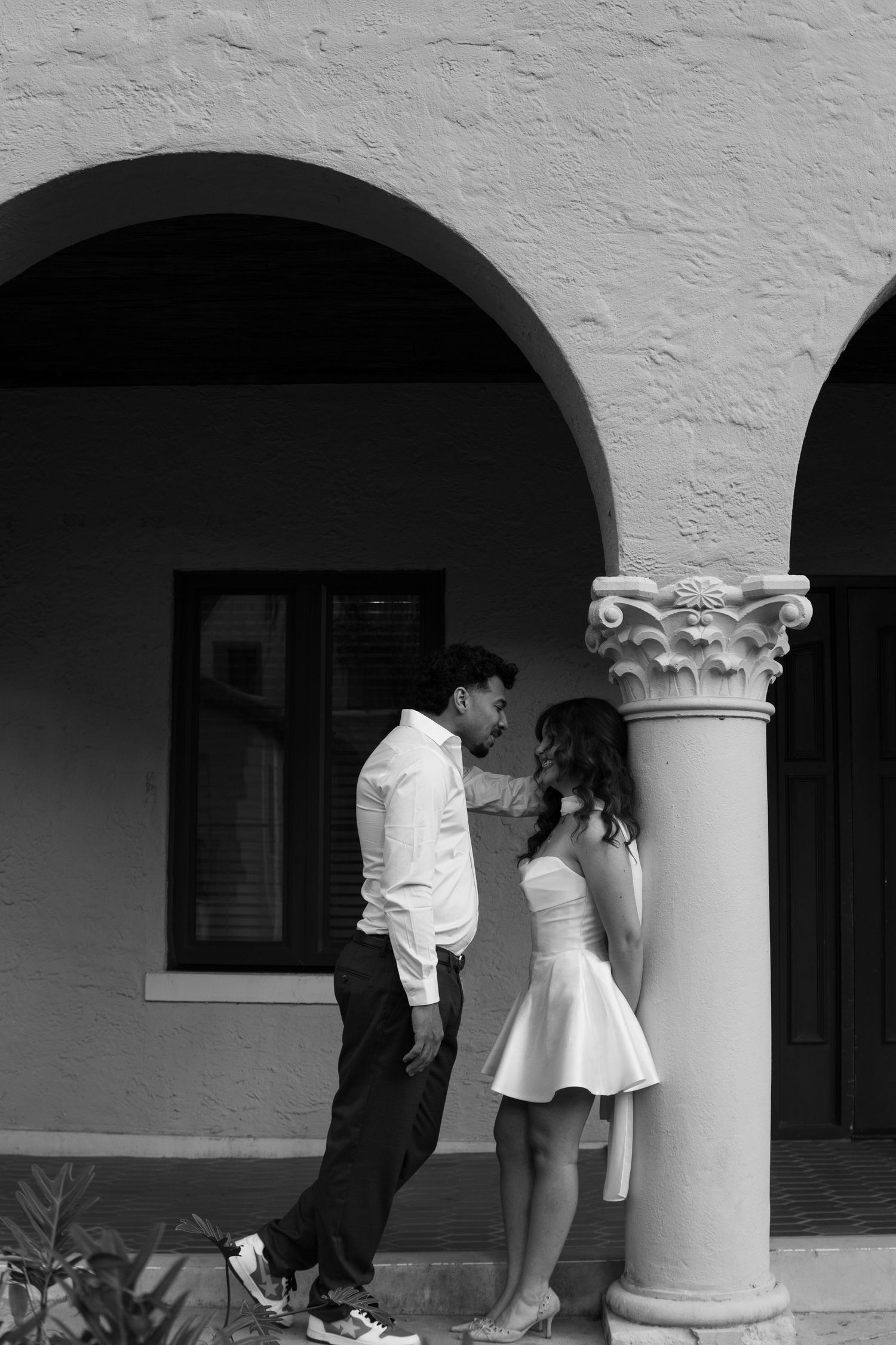 Black and white photo of romantic couple leaning against ornate column under arched building.