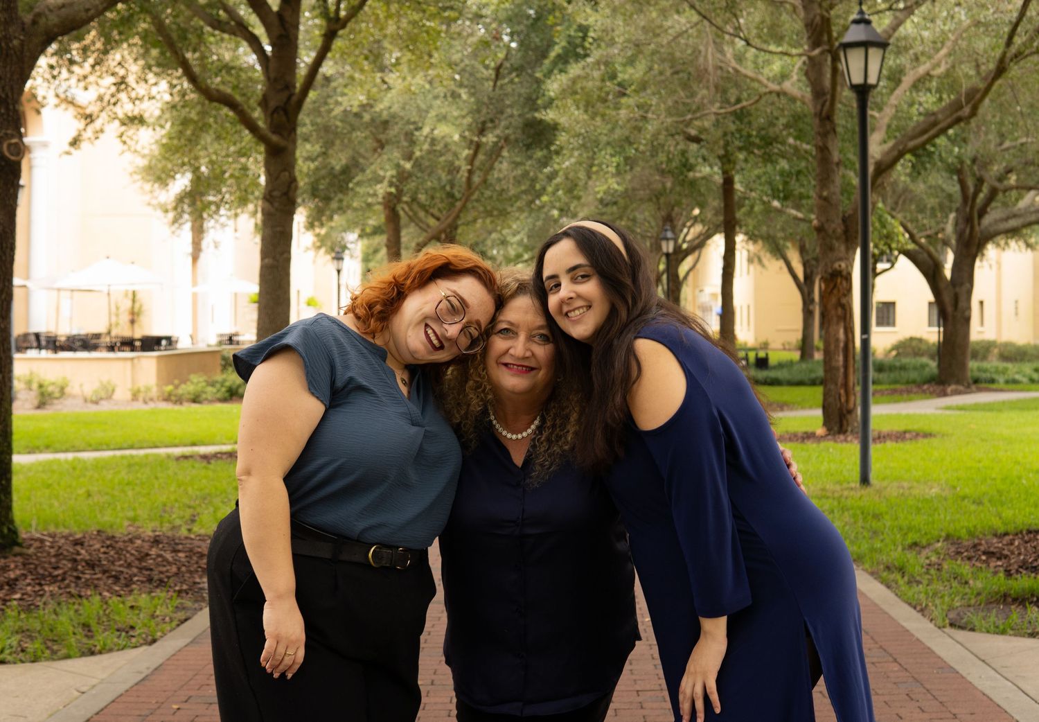 Three women smiling and leaning together outdoors among trees on a brick path.