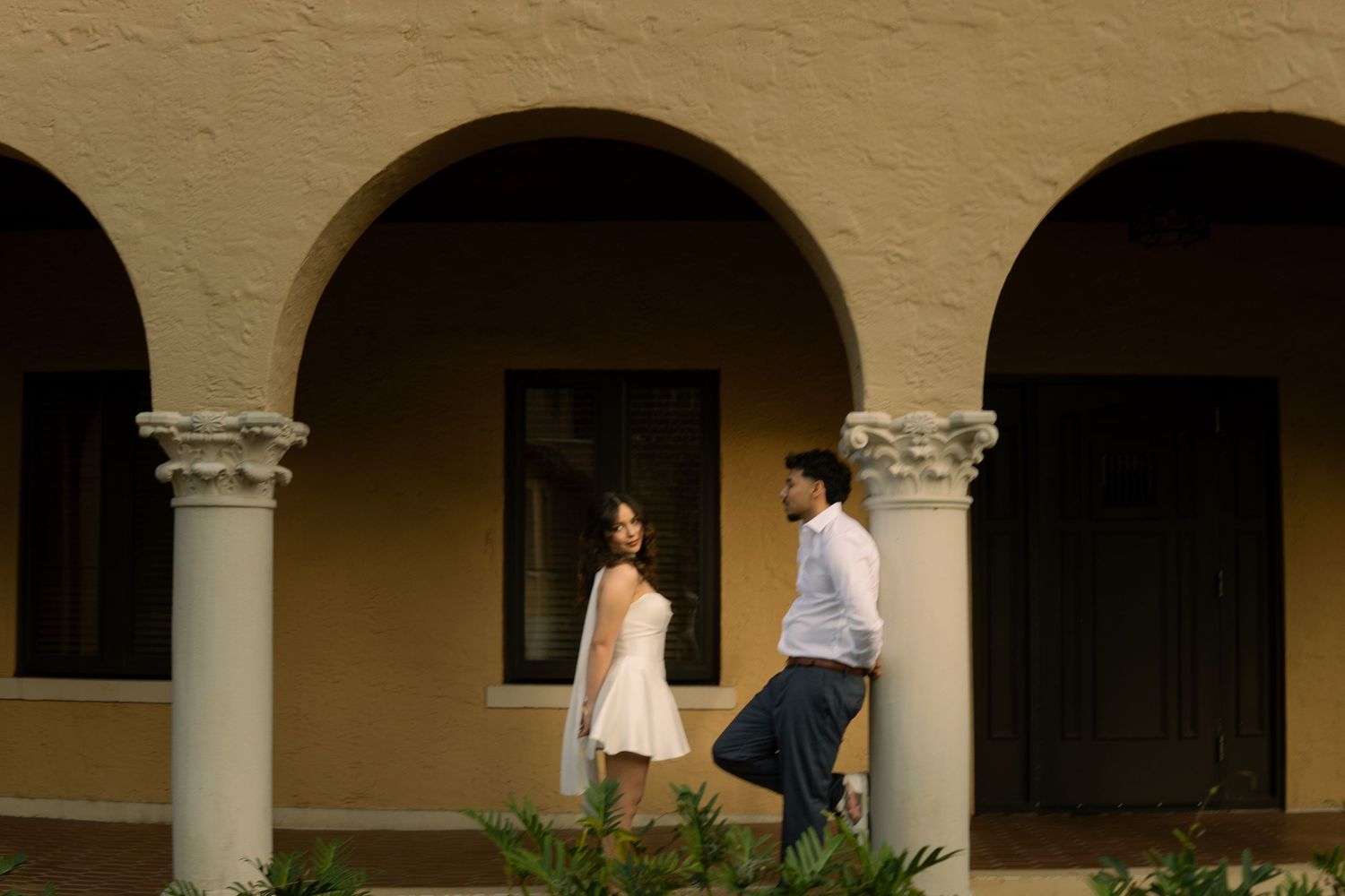 Couple in white dress and casual attire walking through elegant arched colonnade exterior.