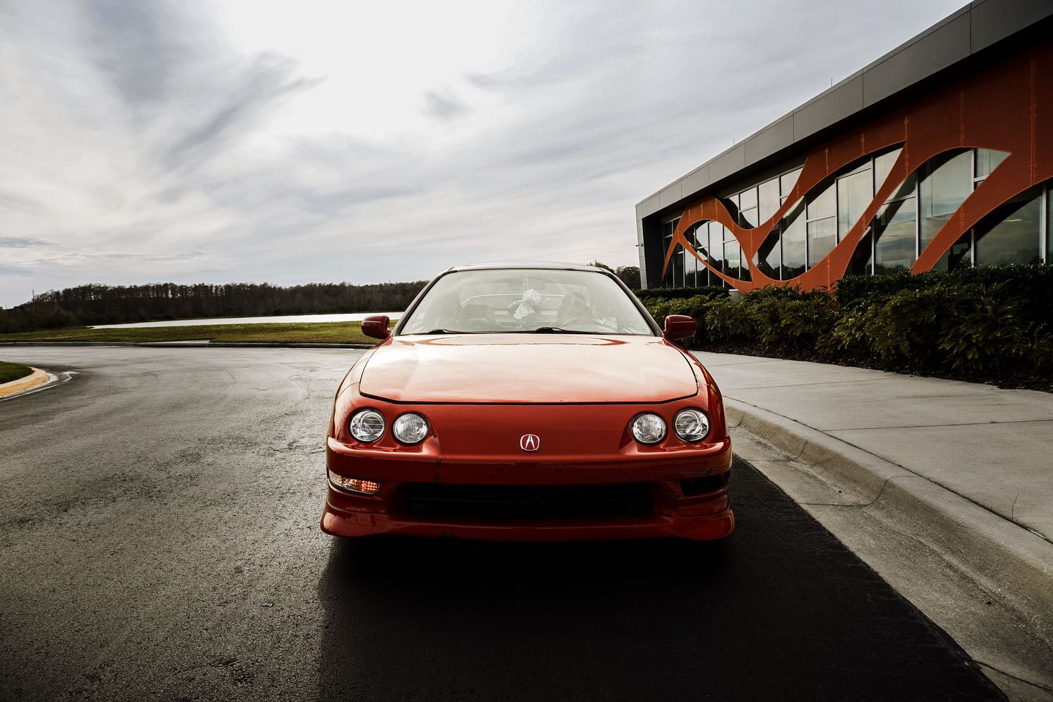 Red Acura Integra coupe photographed head-on in empty parking lot under overcast sky.