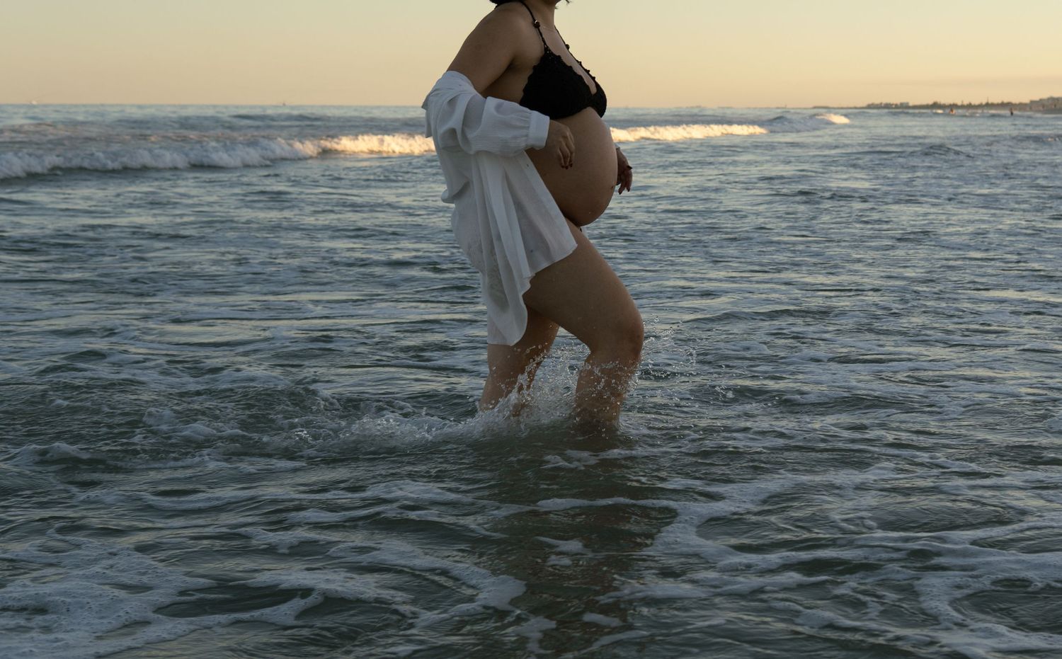 Pregnant woman in black bikini and open white shirt standing in ocean waves at sunset, cradling her baby bump.