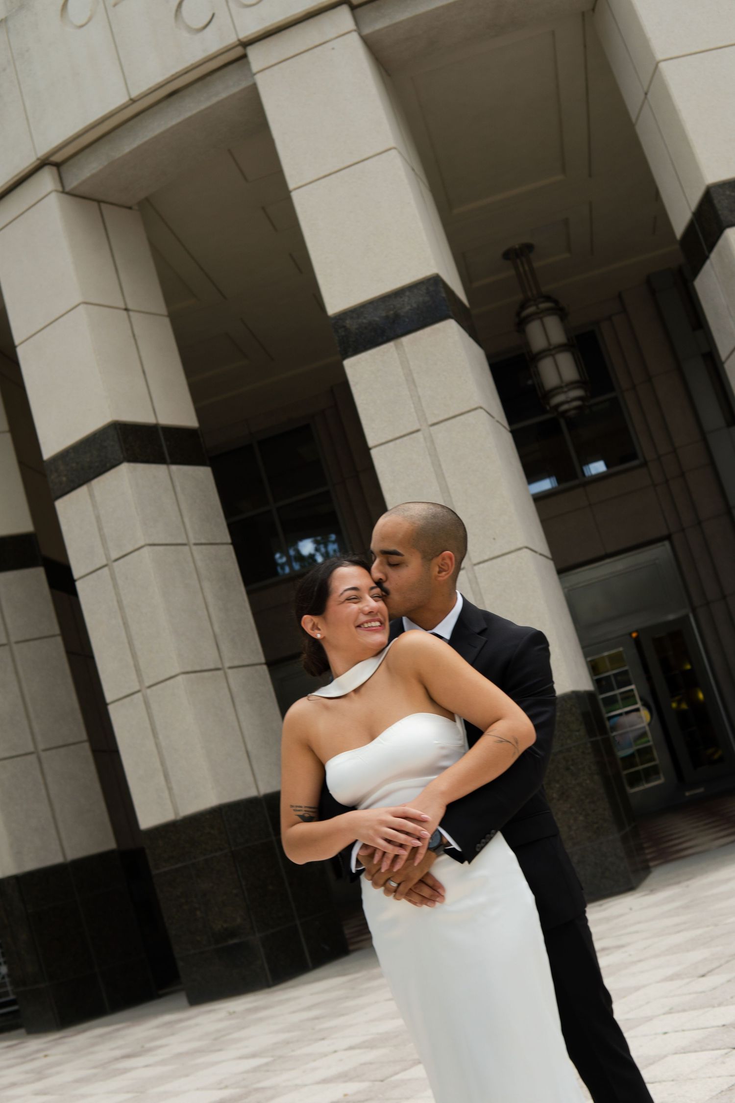 Elegant newlywed couple embracing outside a grand building with large stone columns on their wedding day.