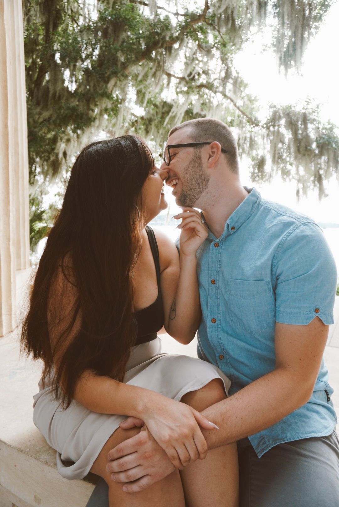 Happy couple shares a romantic moment sitting together under Spanish moss trees near a waterfront.
