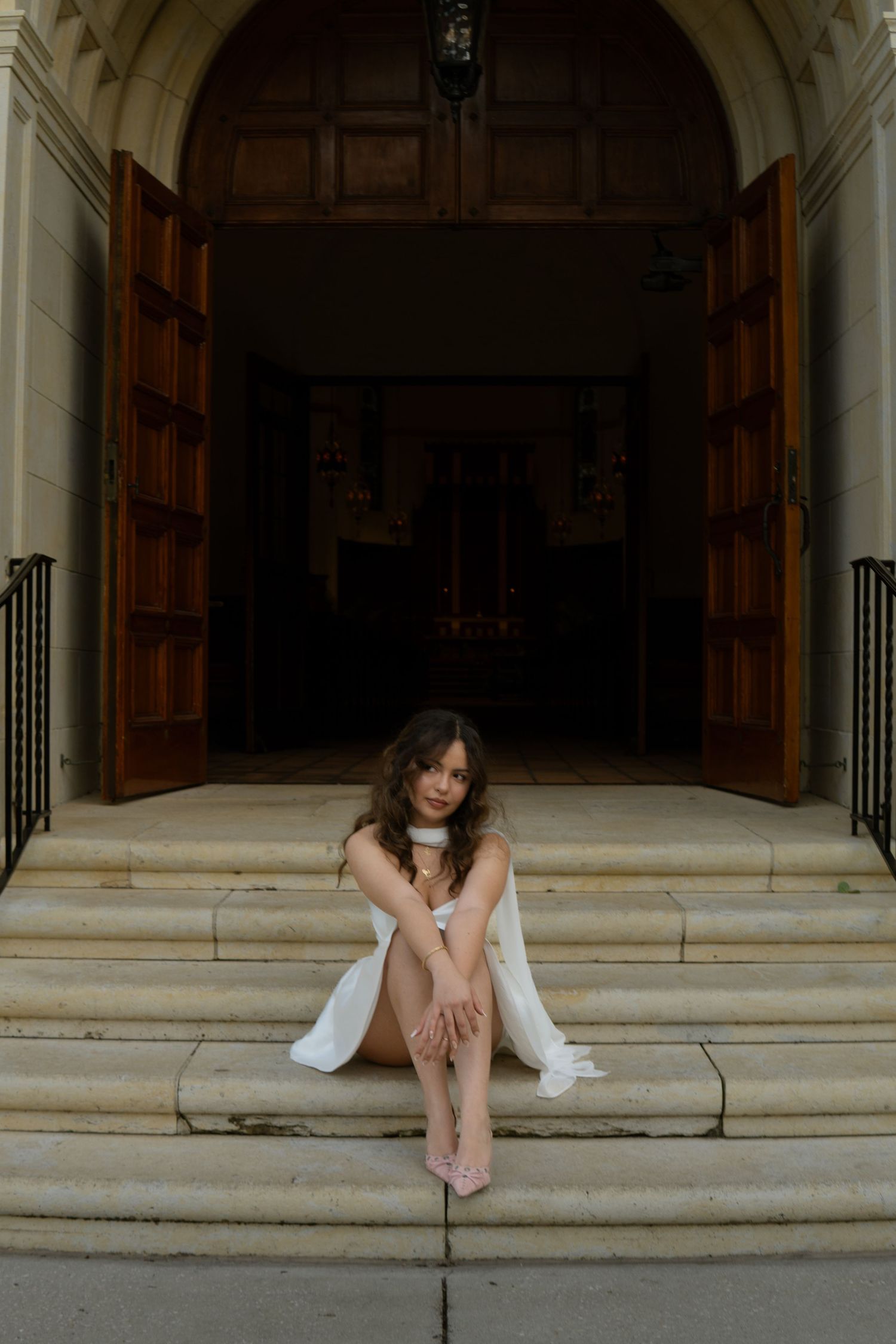 Bride in white dress sitting on stone steps of a grand wooden church entrance.