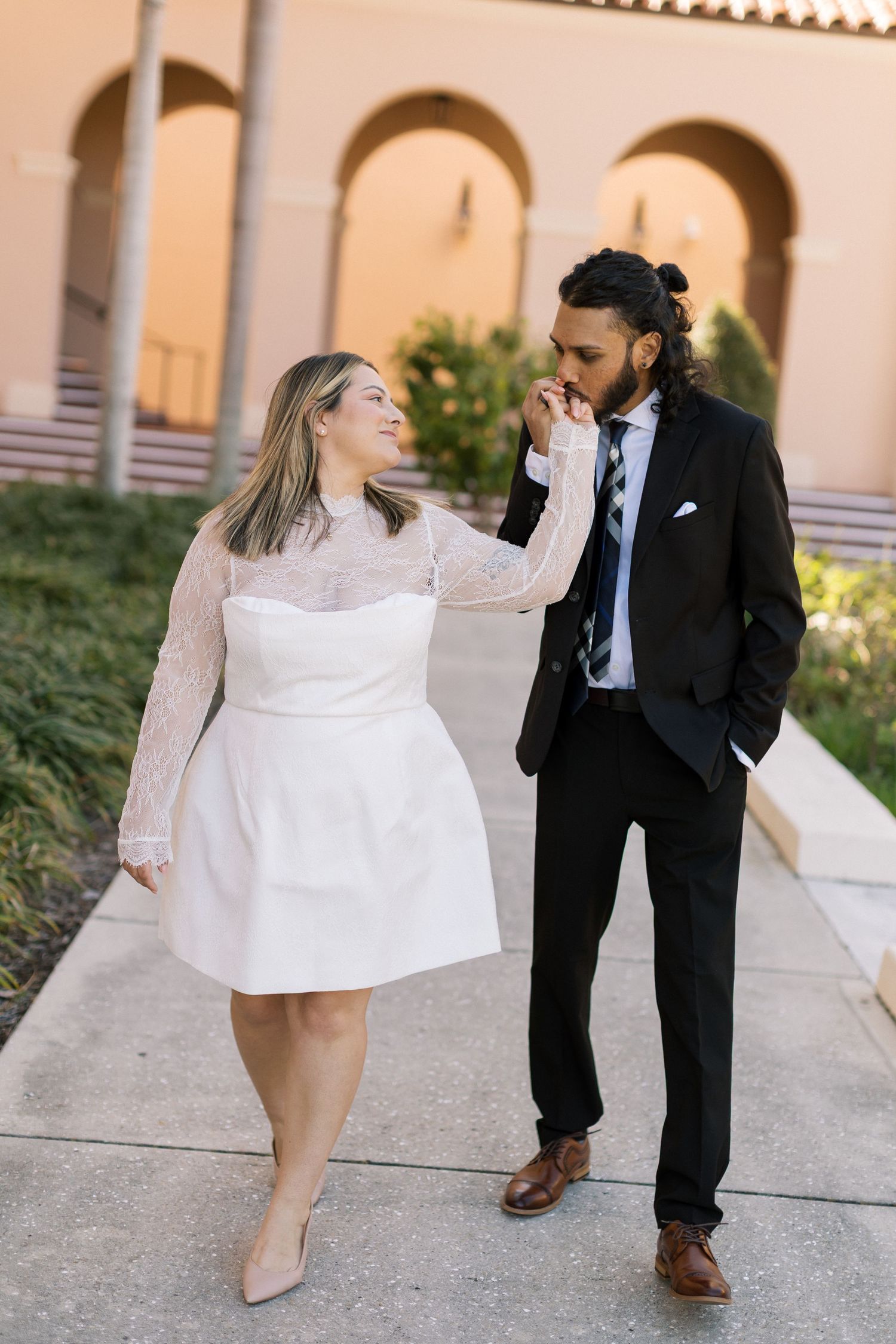 Couple walking outdoors at sunset, woman in white lace mini dress, man in black suit kissing her hand romantically.