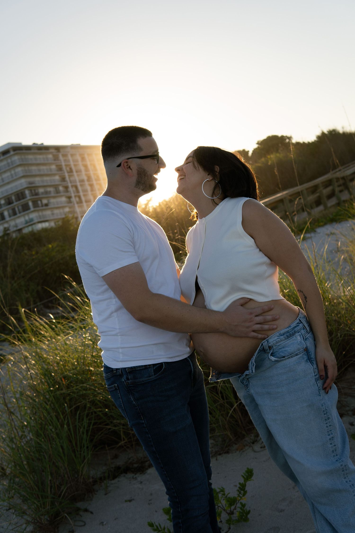 Smiling pregnant couple embracing at golden hour on beach dunes, man in white tee, woman showing bare baby bump.
