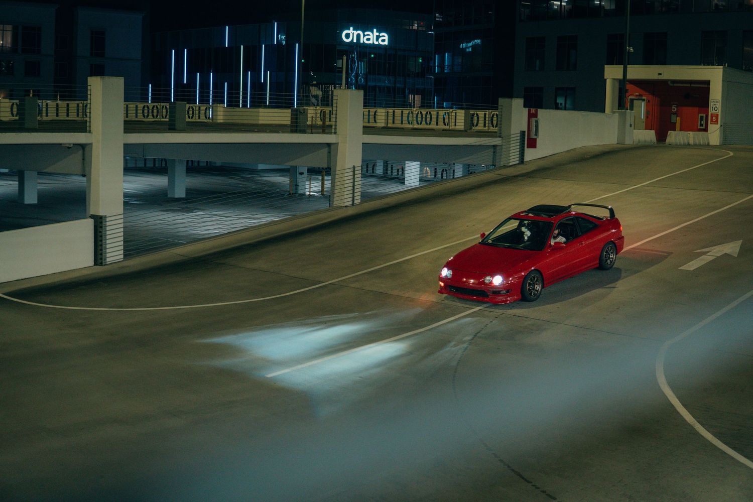 Red Honda Integra driving through an empty parking garage at night with headlights on.