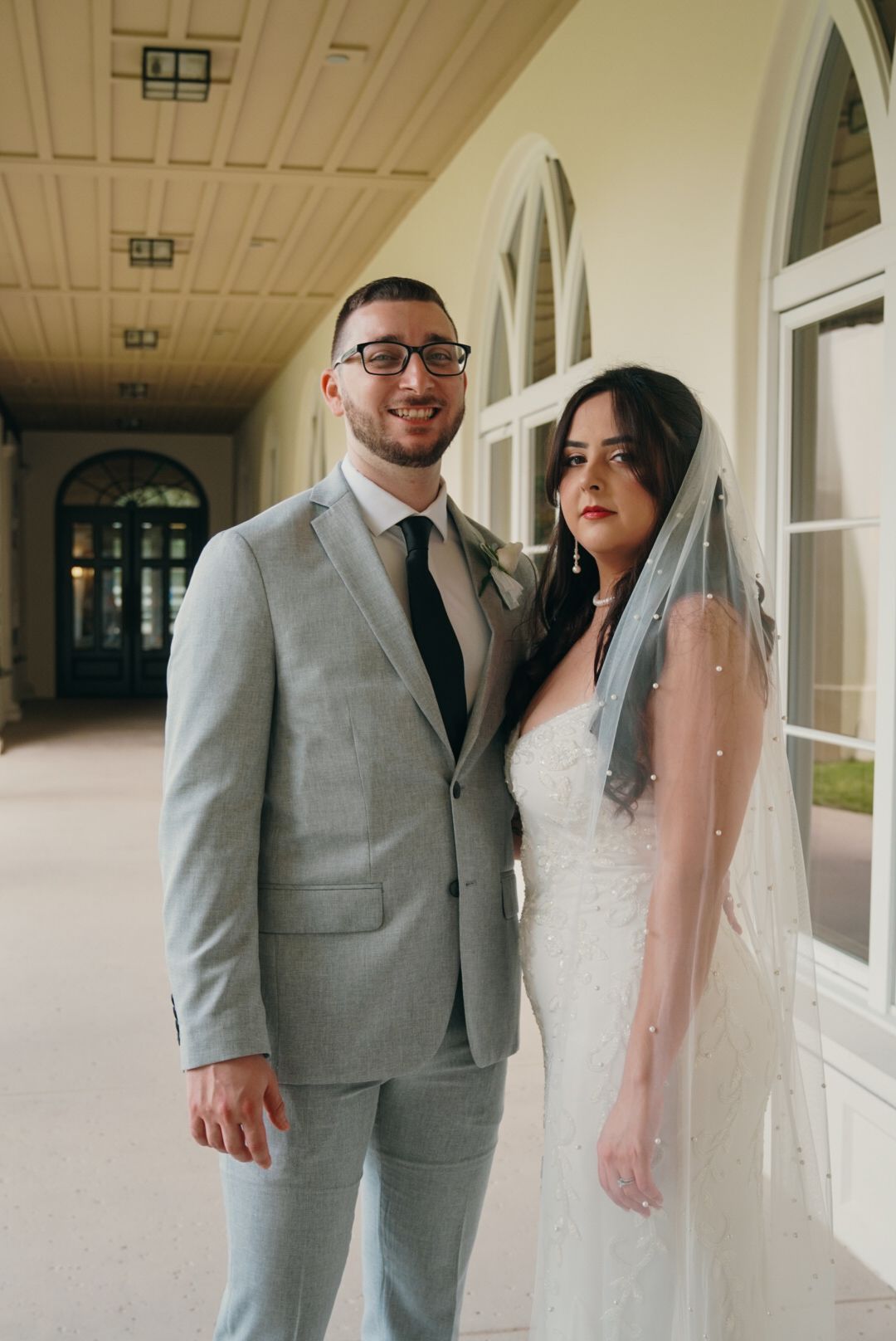 Smiling groom in gray suit with bride in white wedding dress and pearl veil posing in elegant venue hallway.