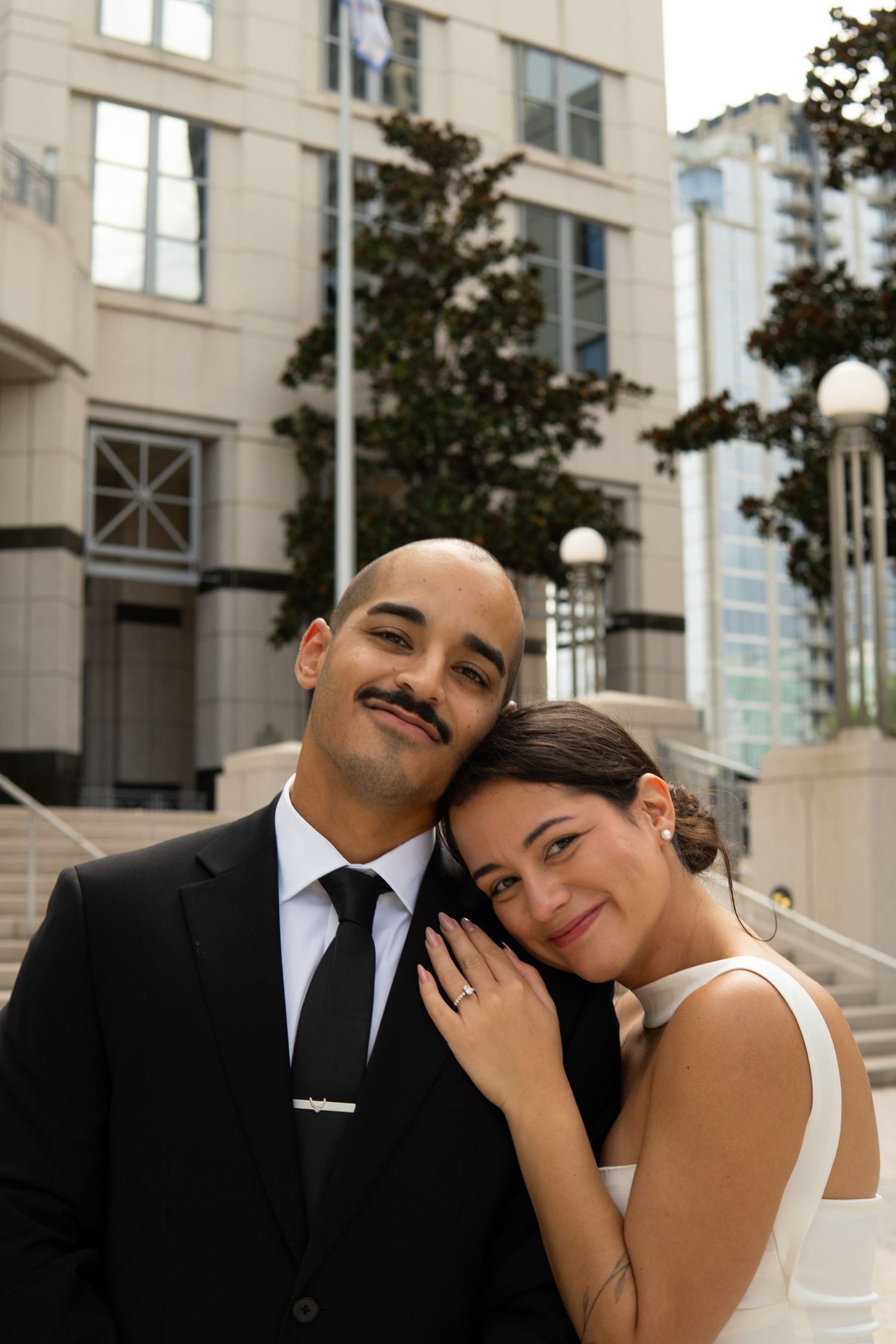Newlywed couple posing together outside, bride resting her head on groom's shoulder, showing engagement ring.