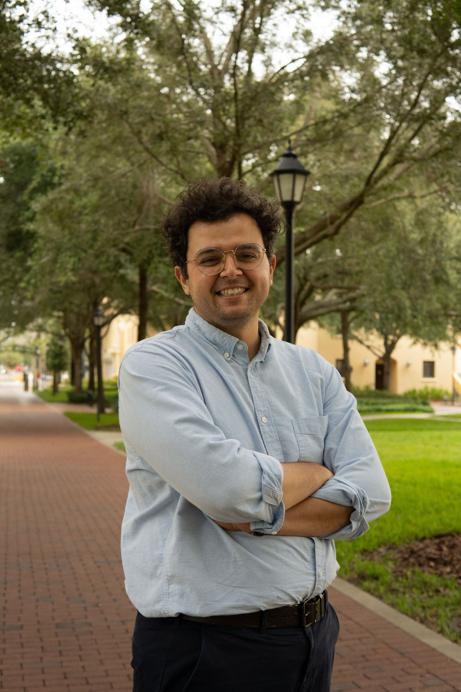 Smiling man with curly hair and glasses standing with arms crossed on a brick walkway.