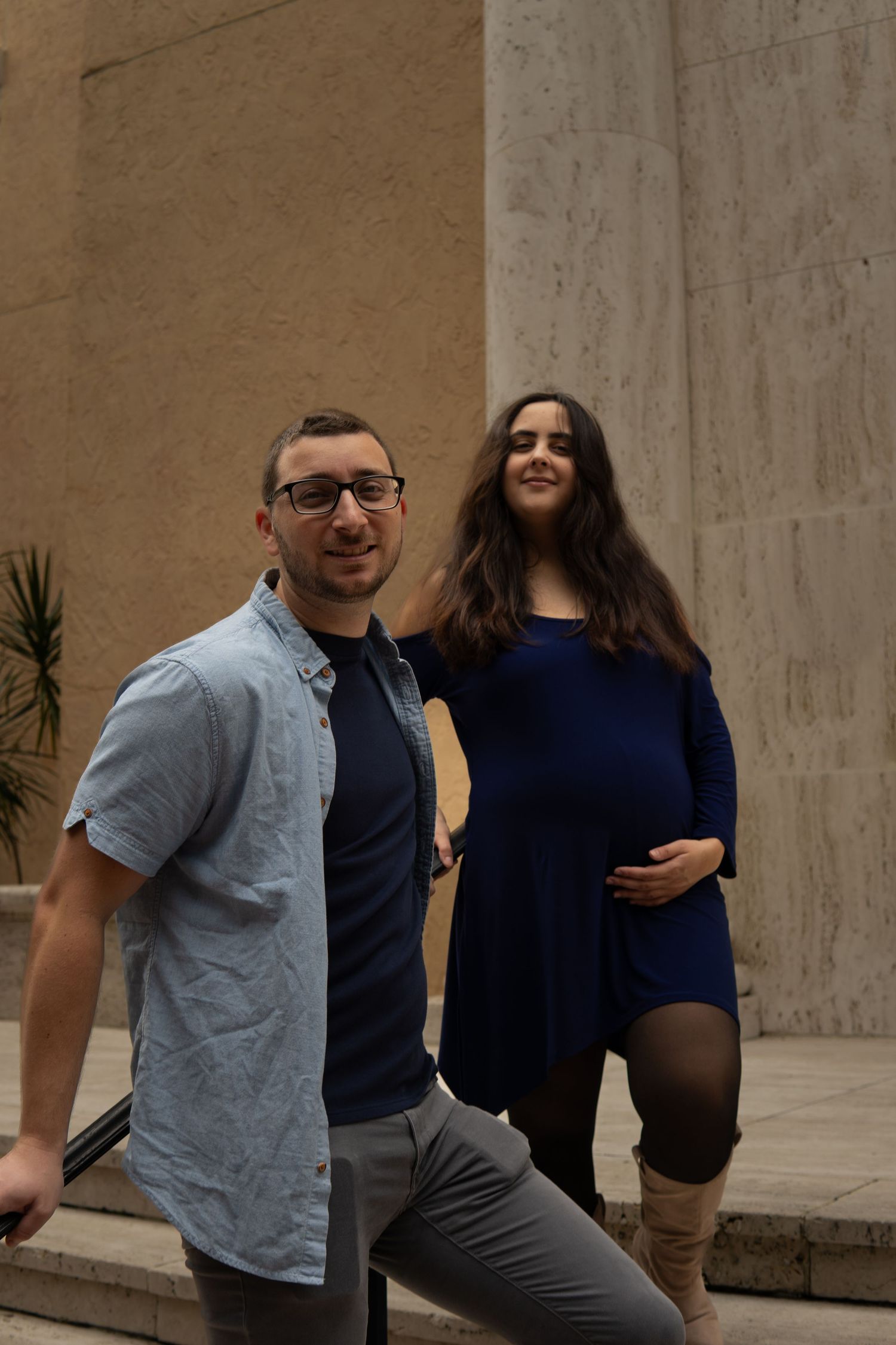 Young man and pregnant woman posing together near tall stone columns outdoors.