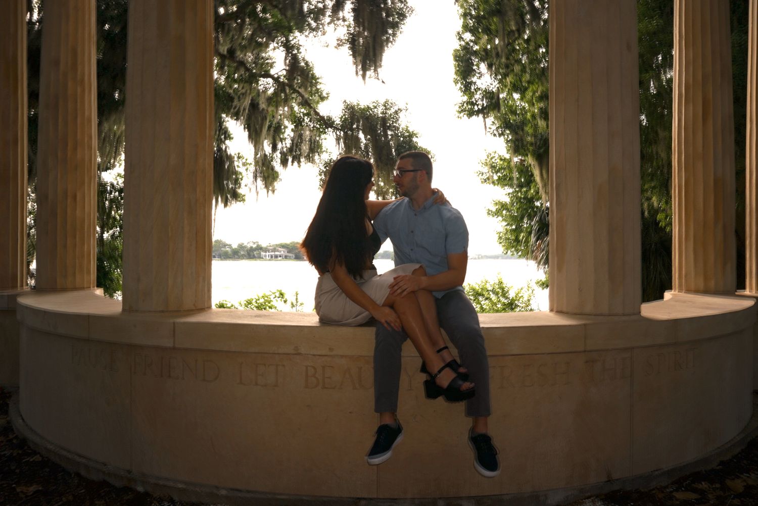 Couple shares a kiss while sitting on a curved marble bench framed by lakeside columns.