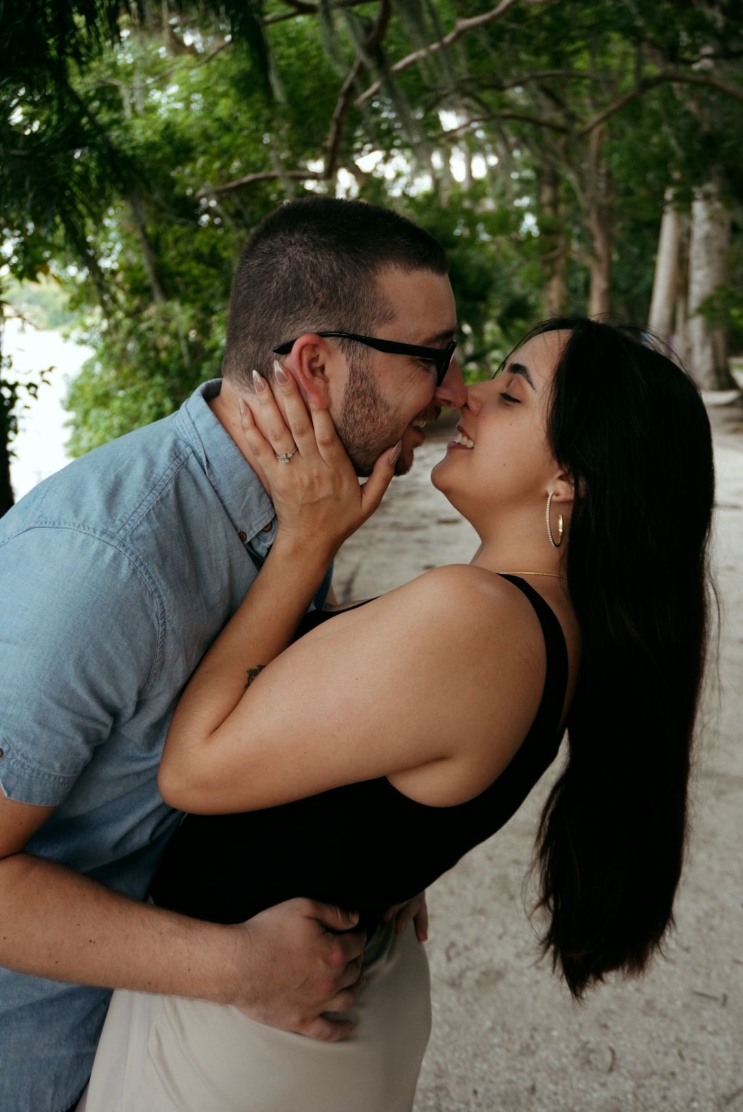 Couple shares a tender kiss outdoors surrounded by lush green trees in a park setting.